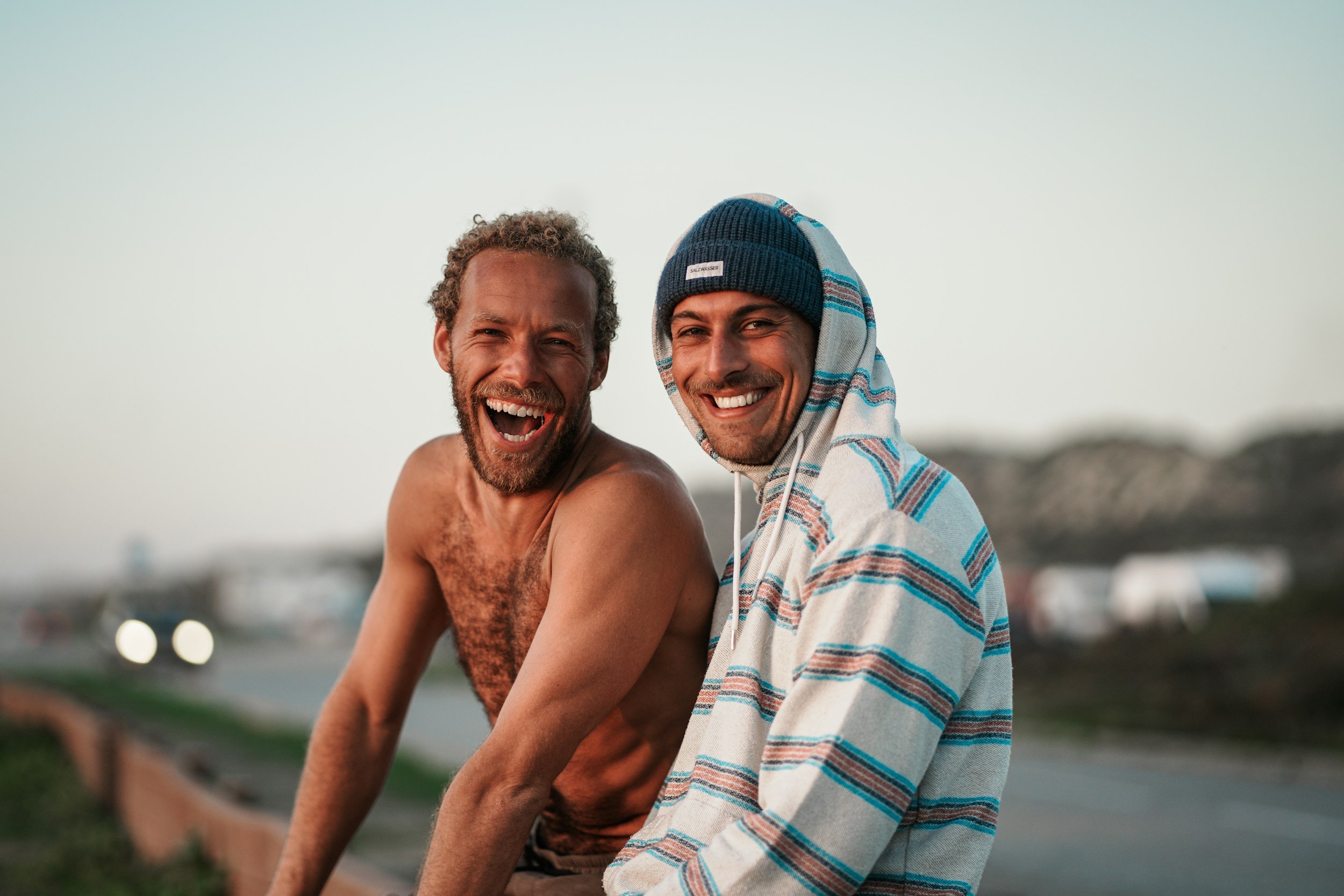 Two men smiling and laughing outdoors on a roadside during daytime. One is shirtless with curly hair and a beard, the other is wearing a striped hoodie and a beanie.