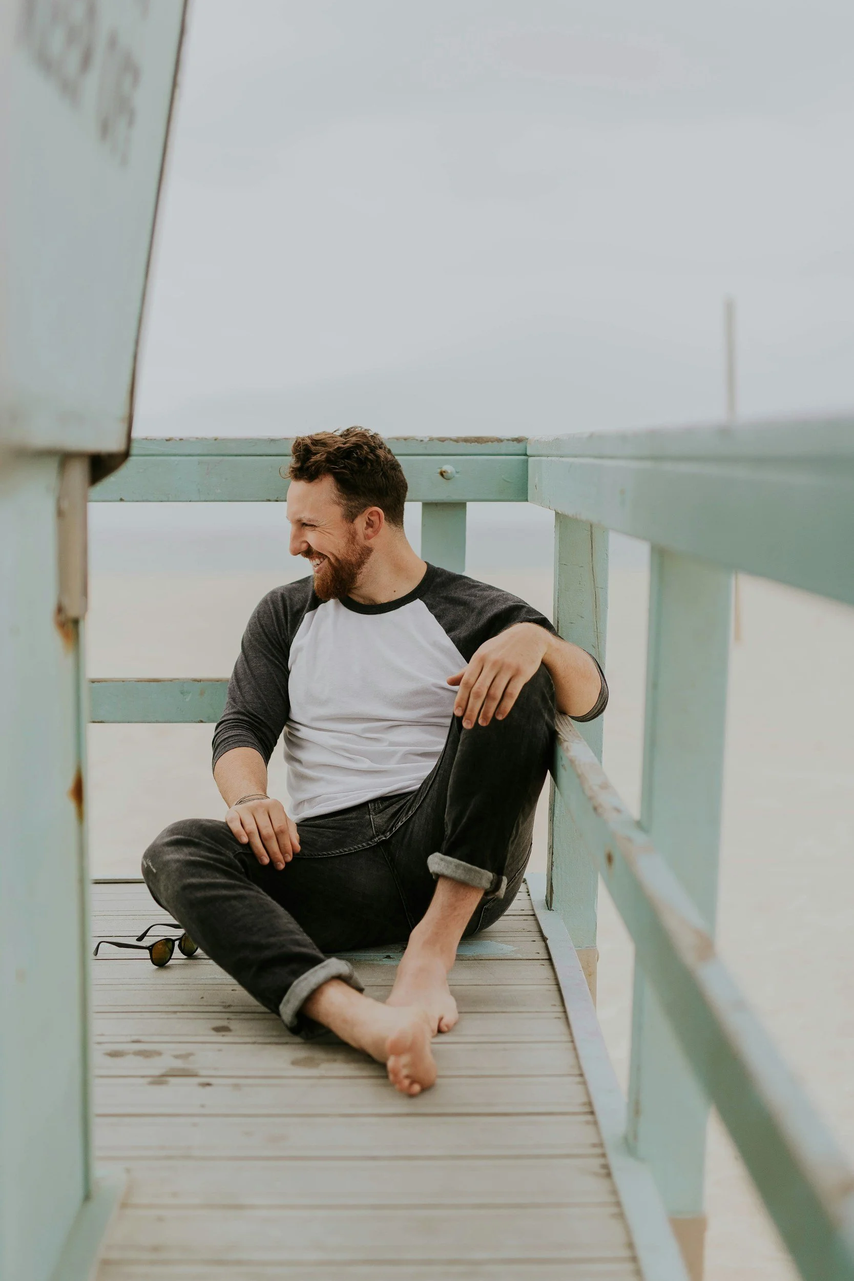 A man with a beard and curly hair sitting barefoot on a wooden deck, smiling and looking to the side, with a pastel-colored railing around him, at what appears to be a beach or lakeside location.