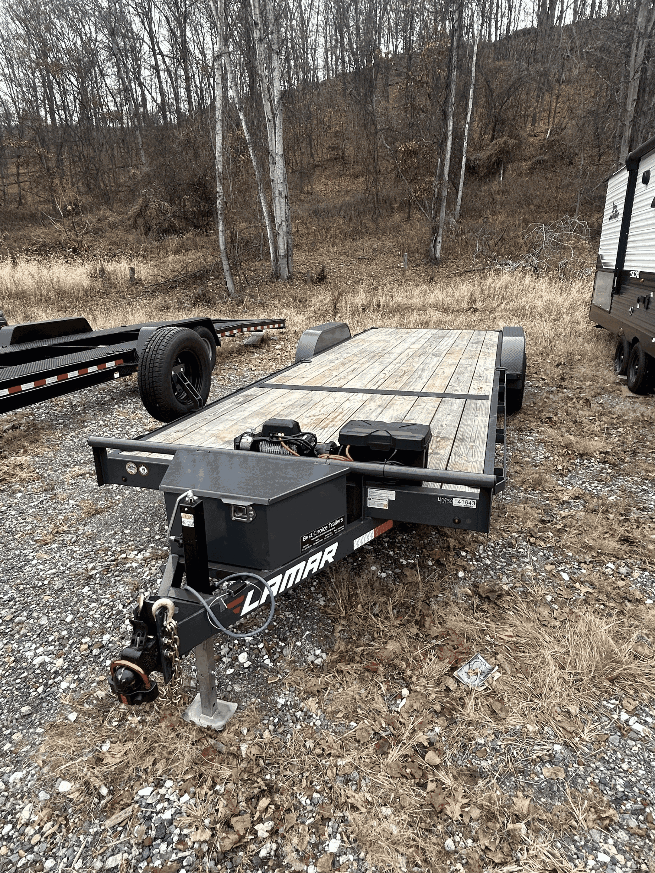 A flatbed trailer with a wooden deck parked on a gravel area with dry grass and trees in the background.