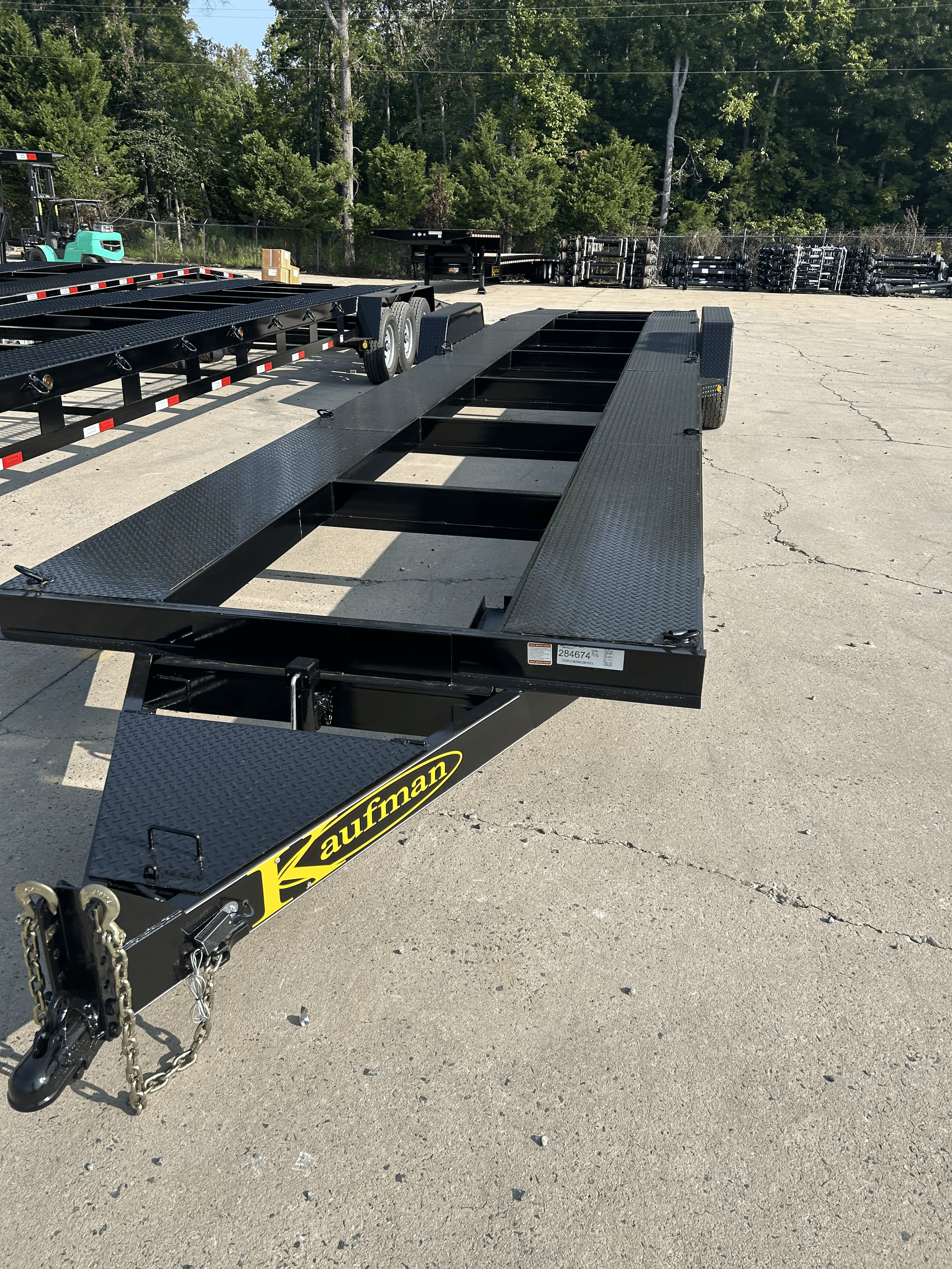 Black Kaufman flatbed trailer with no bed, parked on a cracked asphalt lot, with trees and other trailers in the background.