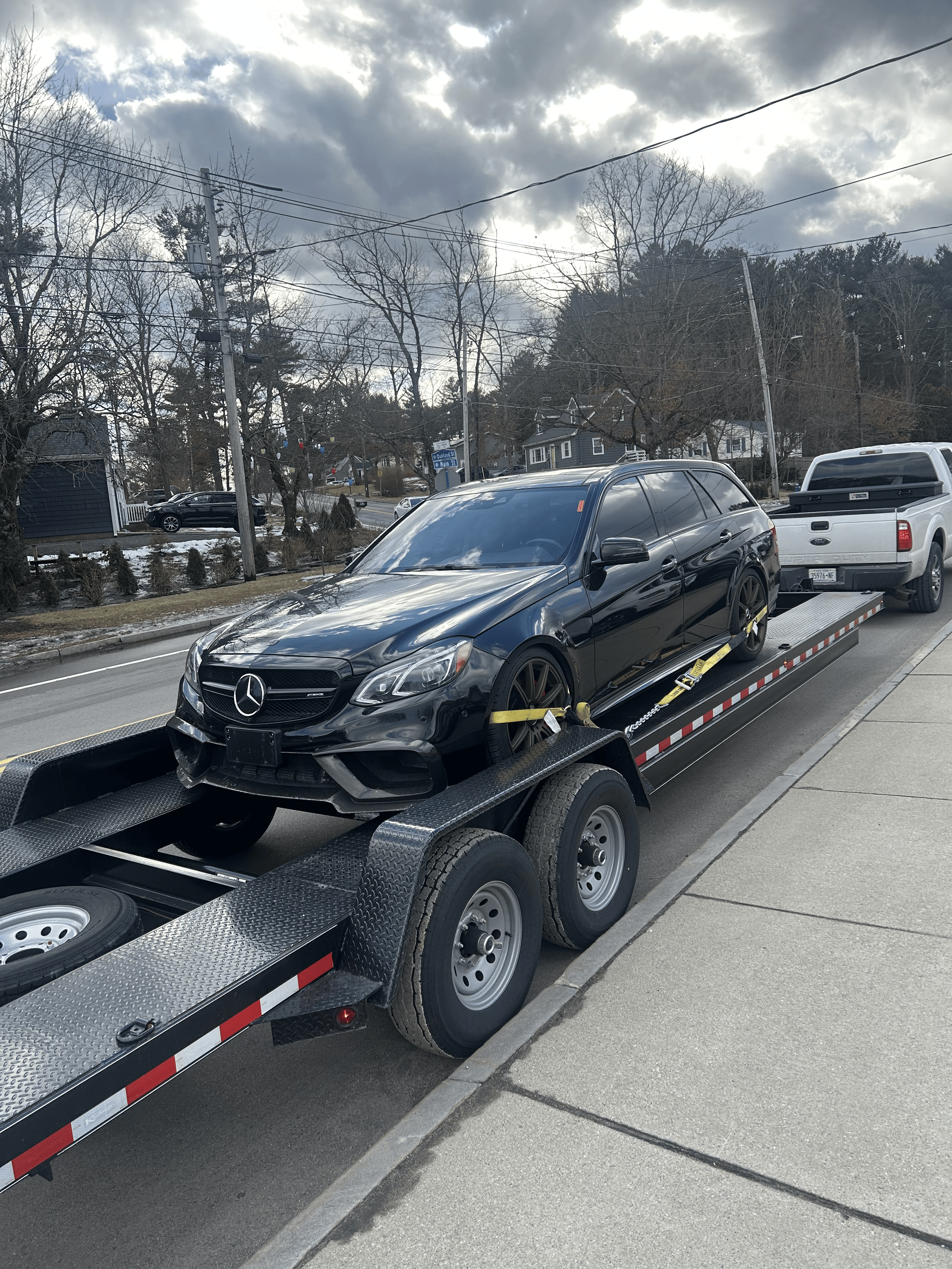 A black Mercedes-Benz car secured on a flatbed tow truck on a city street with overcast weather and some houses and trees in the background.