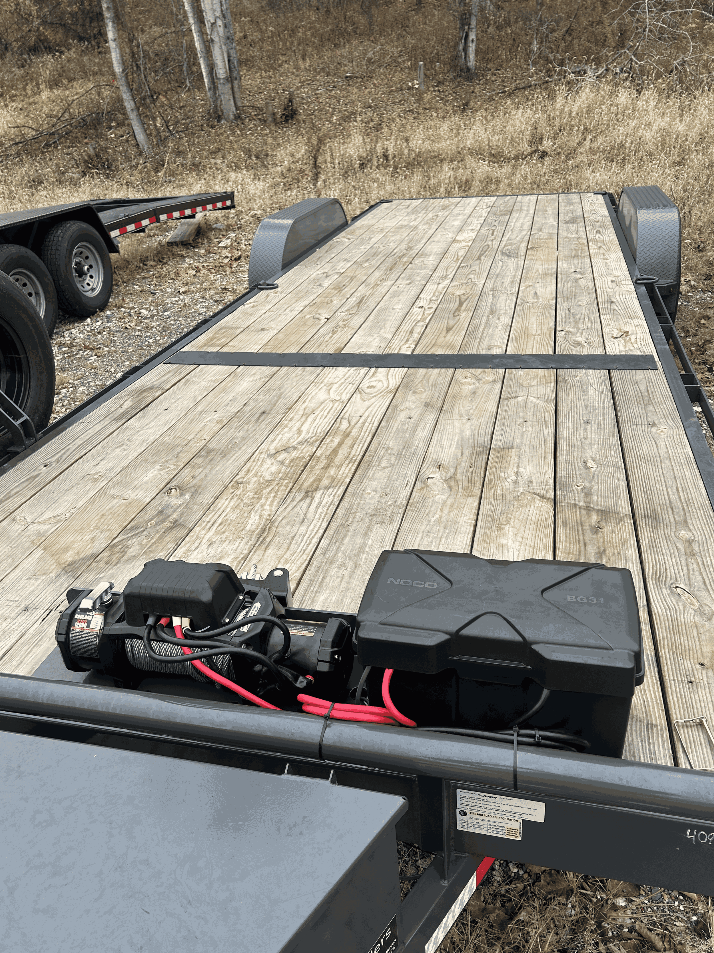 Empty flatbed trailer with wooden planks and a black battery pack on the front, parked on a dirt lot with dried grass and bare trees in the background.