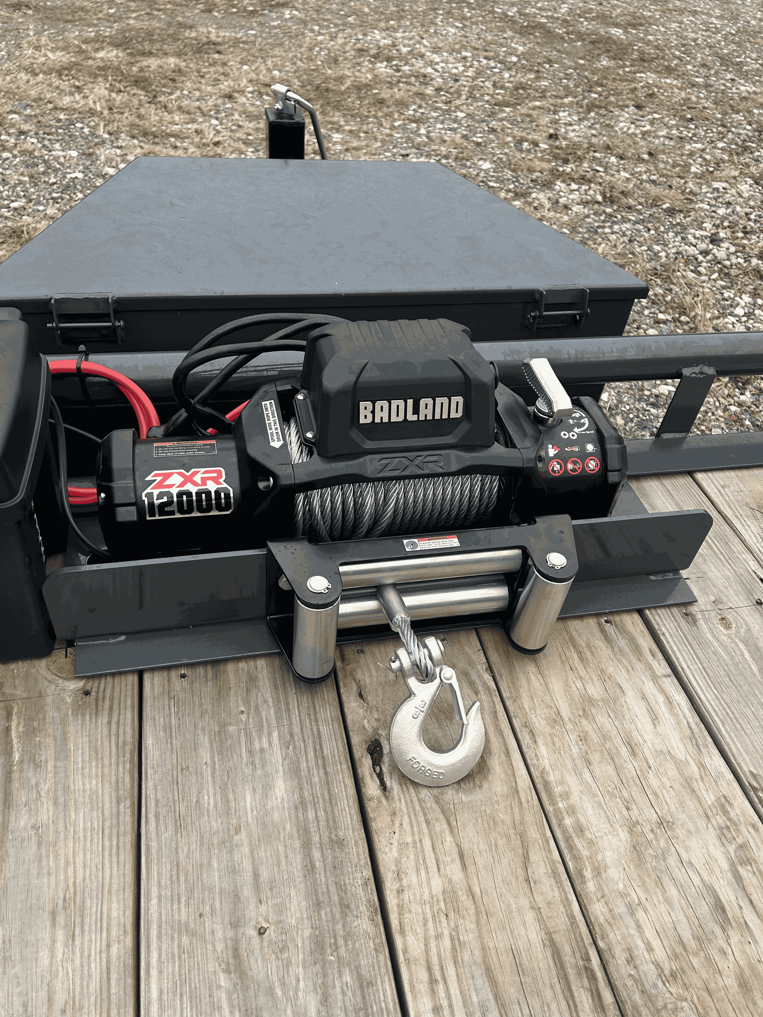 Close-up of a heavy-duty electric winch mounted on a wooden deck, with a metal hook hanging from the cable, set outdoors on a gravel surface.
