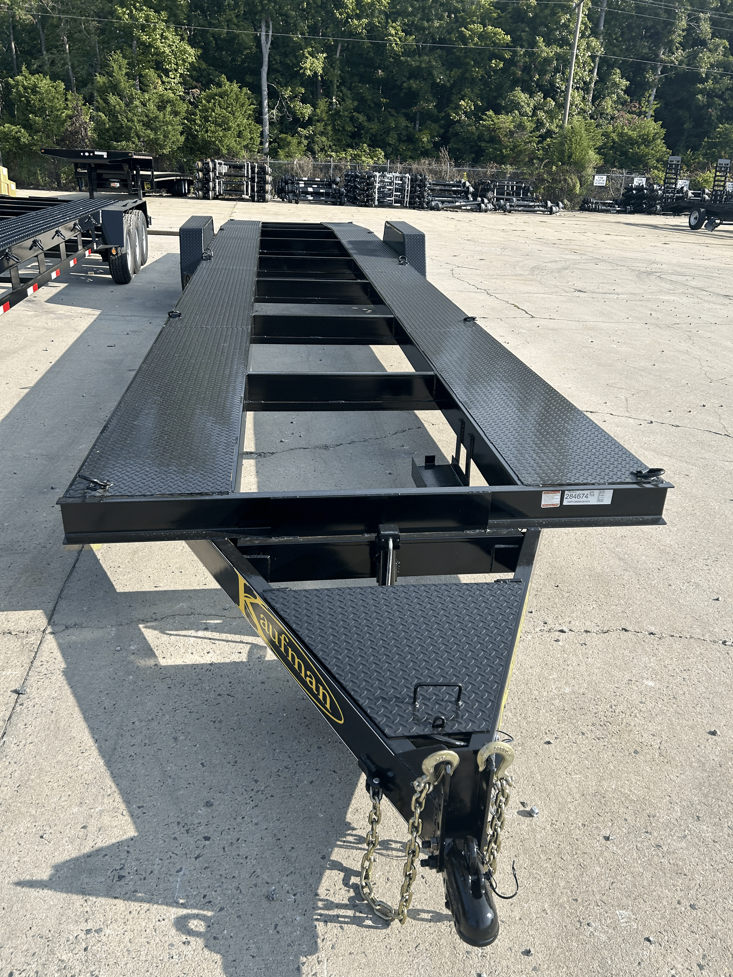 A black Kaufman heavy-duty flatbed utility trailer stands on a lot with other trailers and equipment, with a background of green trees and a chain-link fence.