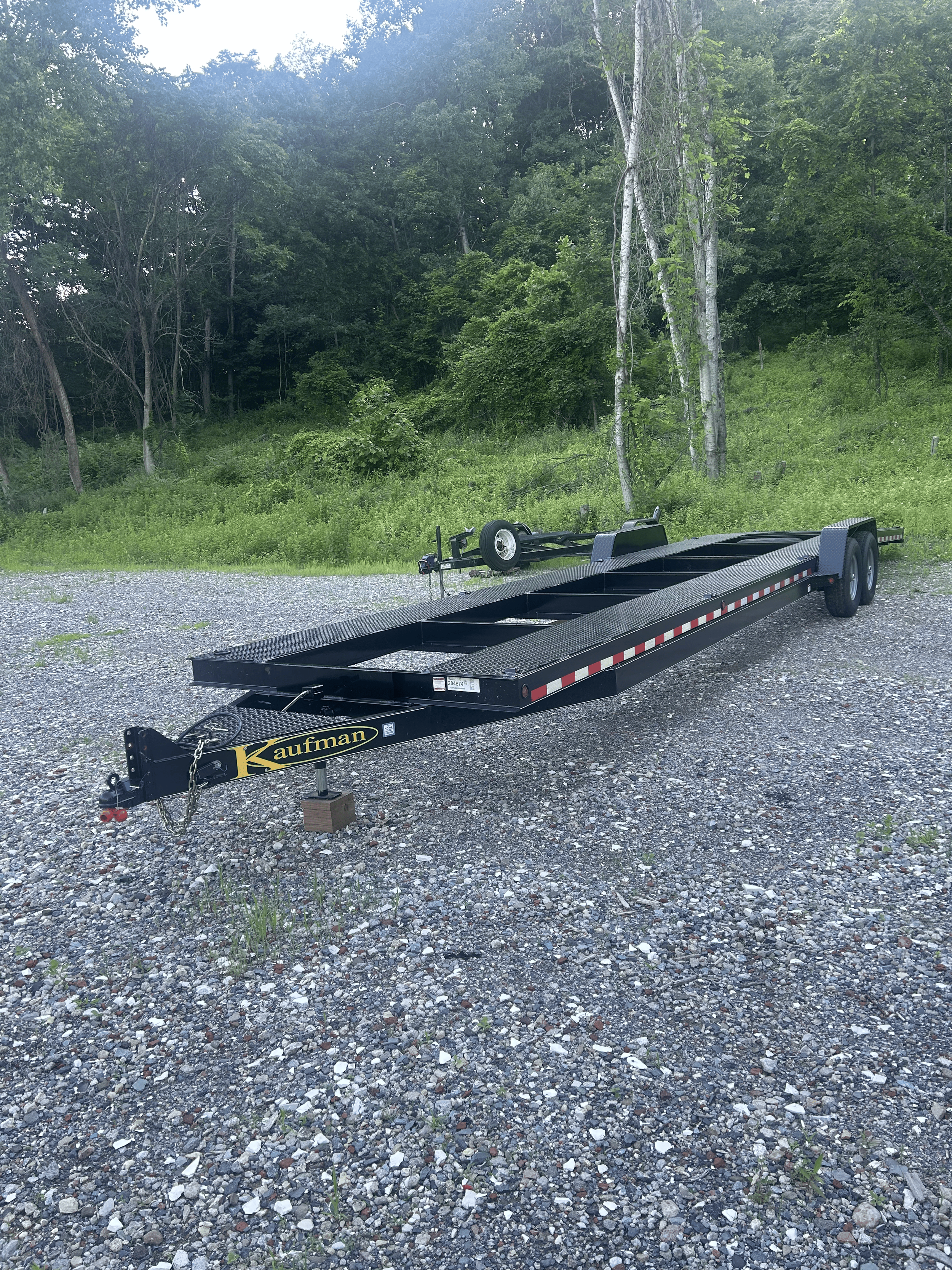 Black flatbed trailer with dual axles parked on gravel ground, surrounded by trees and greenery.