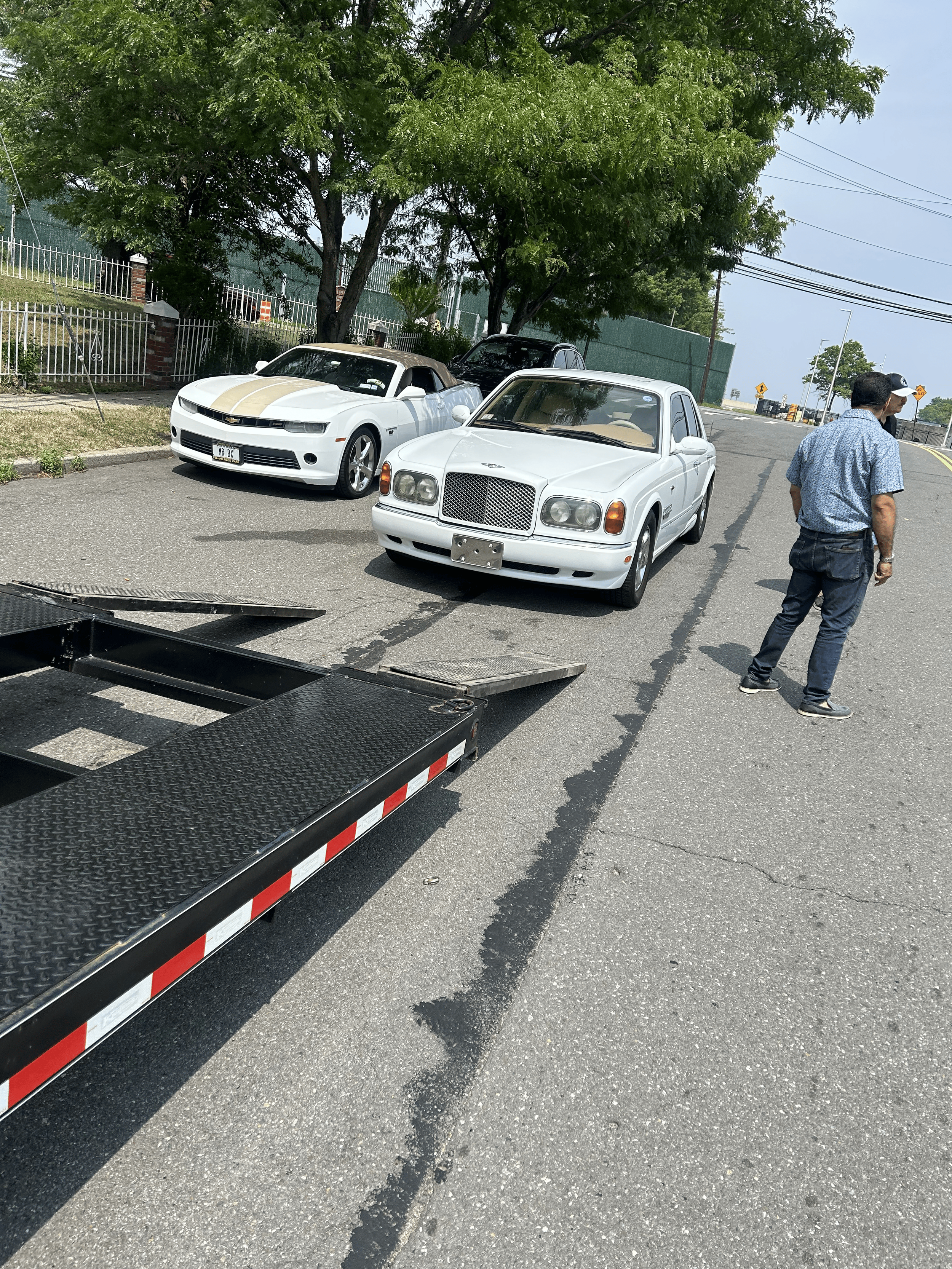 Two parked cars, a white sports car with black and gold stripes and a white luxury sedan, on a street with a person walking nearby, a tow truck in the foreground, trees and a fence in the background.