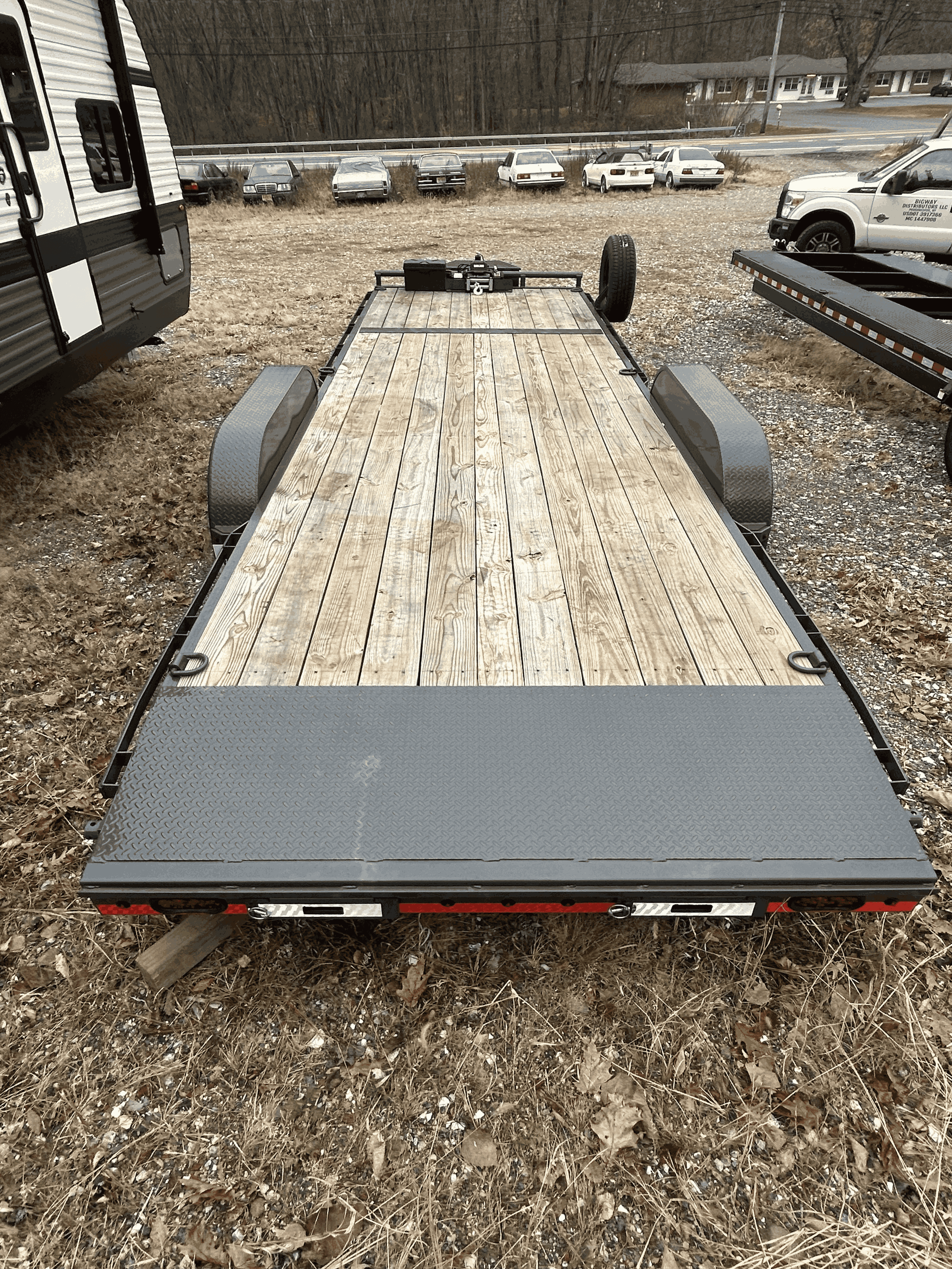 A flatbed trailer with a wooden deck parked on a dirt and gravel lot, surrounded by other vehicles including cars and trucks, under a cloudy sky.