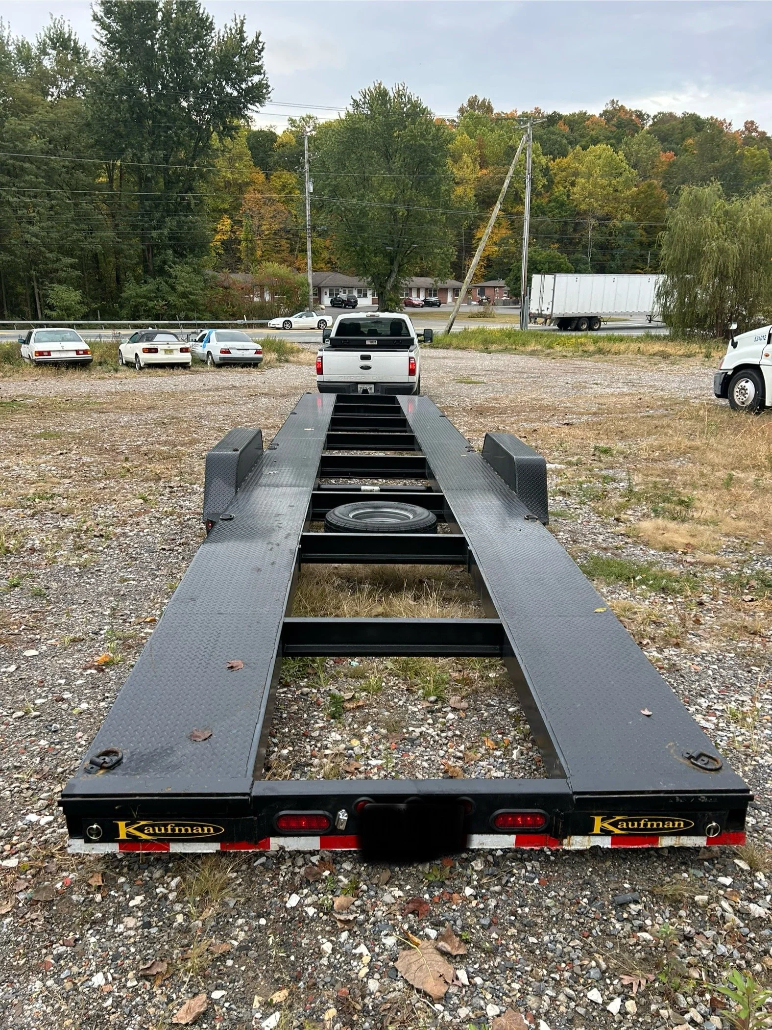 A black flatbed trailer with a spare tire on its frame, parked outdoors on a gravel lot, with multiple cars and trees in the background.