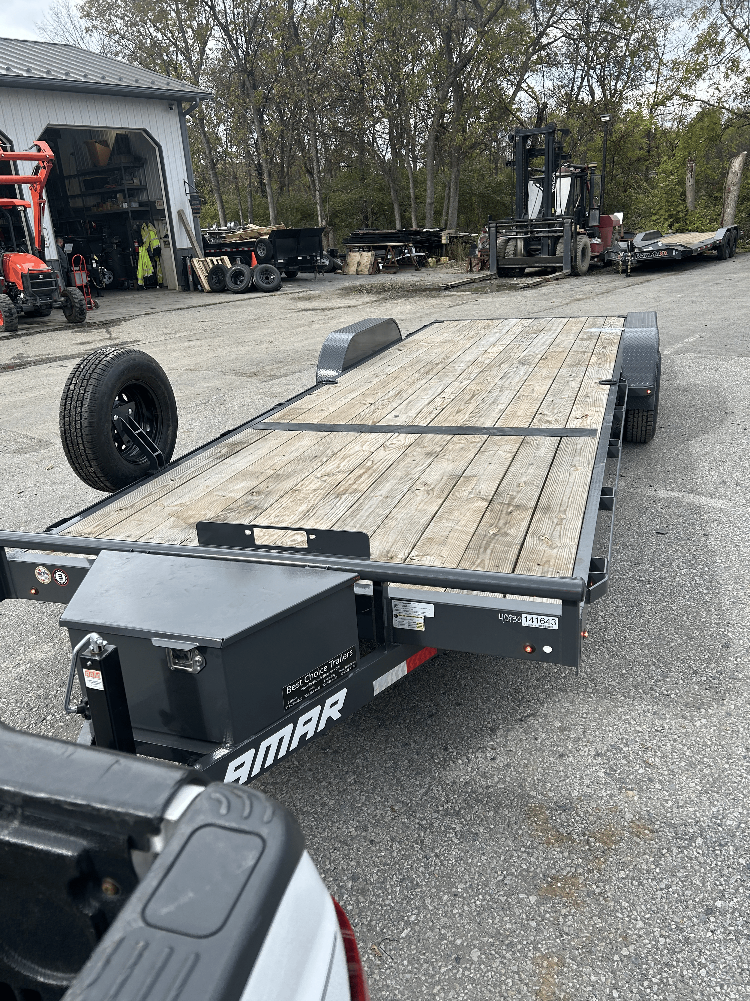 Flatbed trailer with a wooden deck parked on a gravel lot near a garage and industrial equipment.