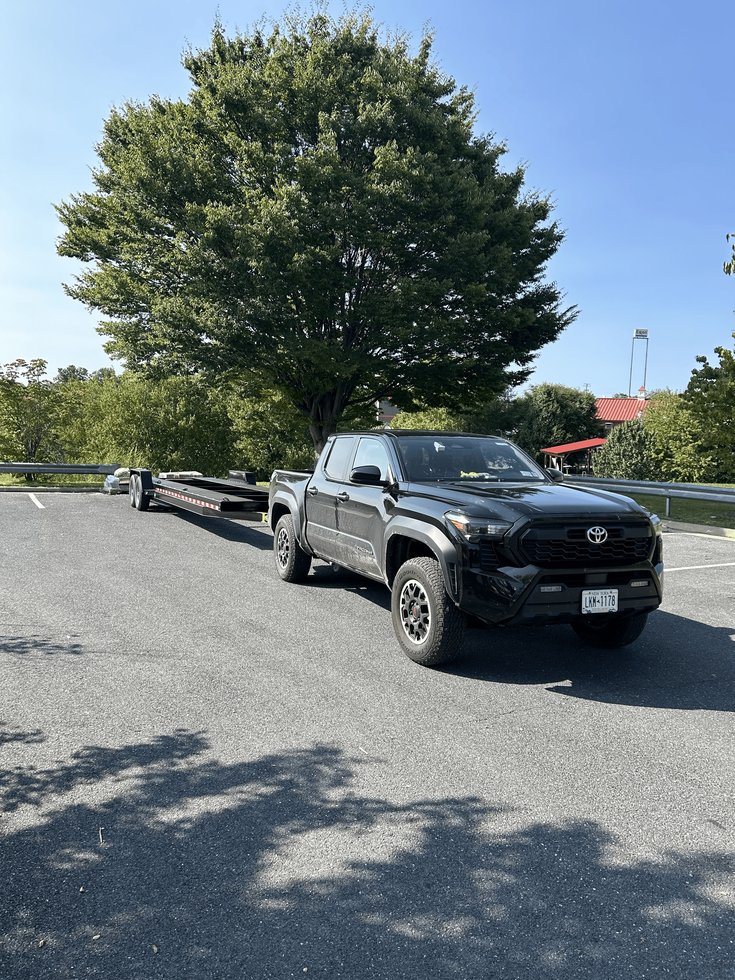 A black Toyota pickup truck with a trailer attached, parked in a parking lot under a large green tree on a sunny day.
