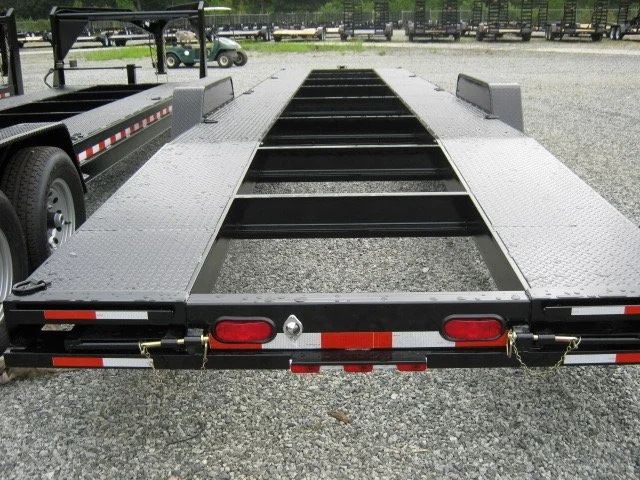 Empty flatbed trailer with a metal frame and black rollers, parked on gravel with a golf cart and benches in the background.