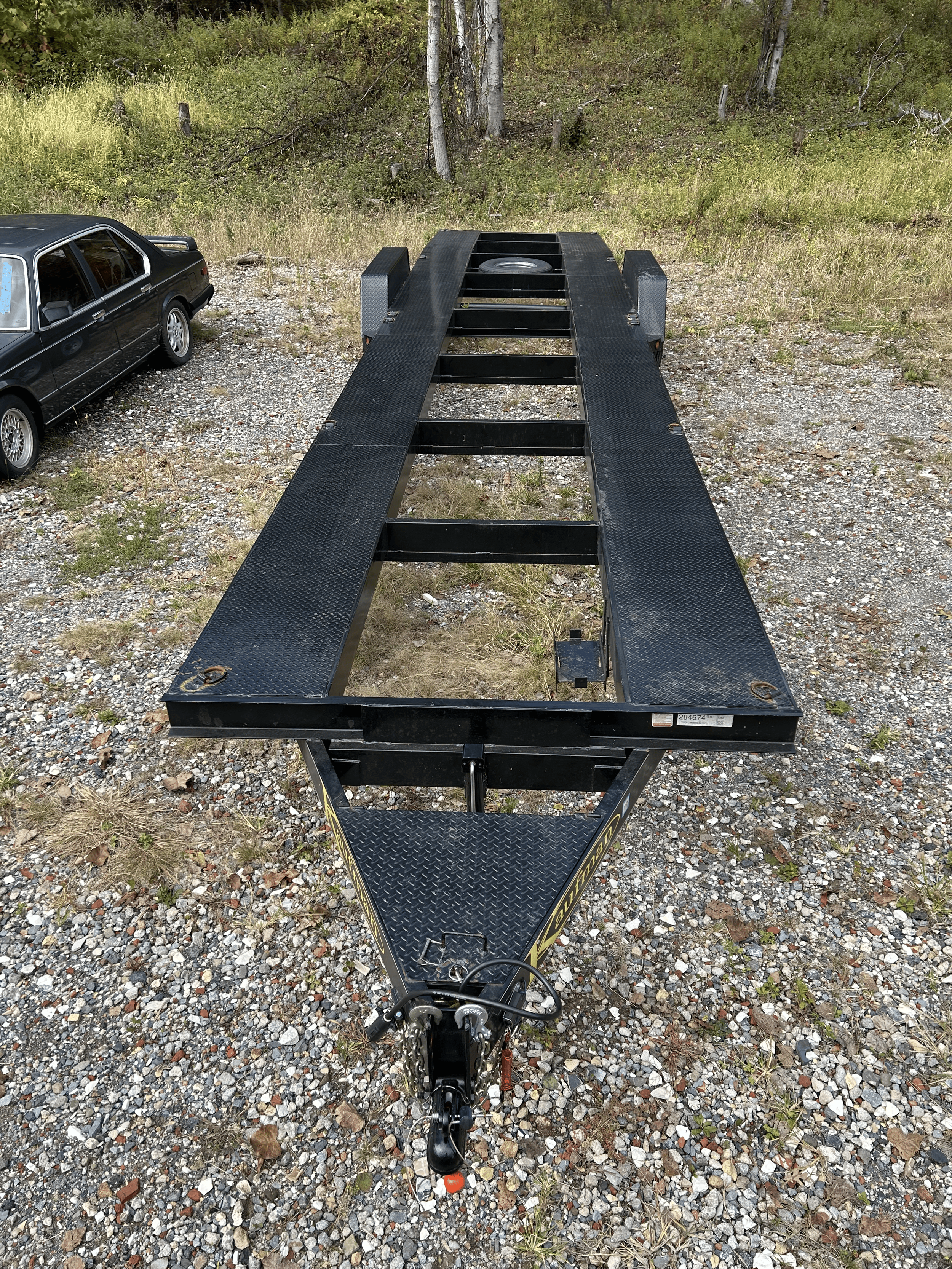A black flatbed car trailer parked on a gravel surface with a black car parked nearby, and trees in the background.