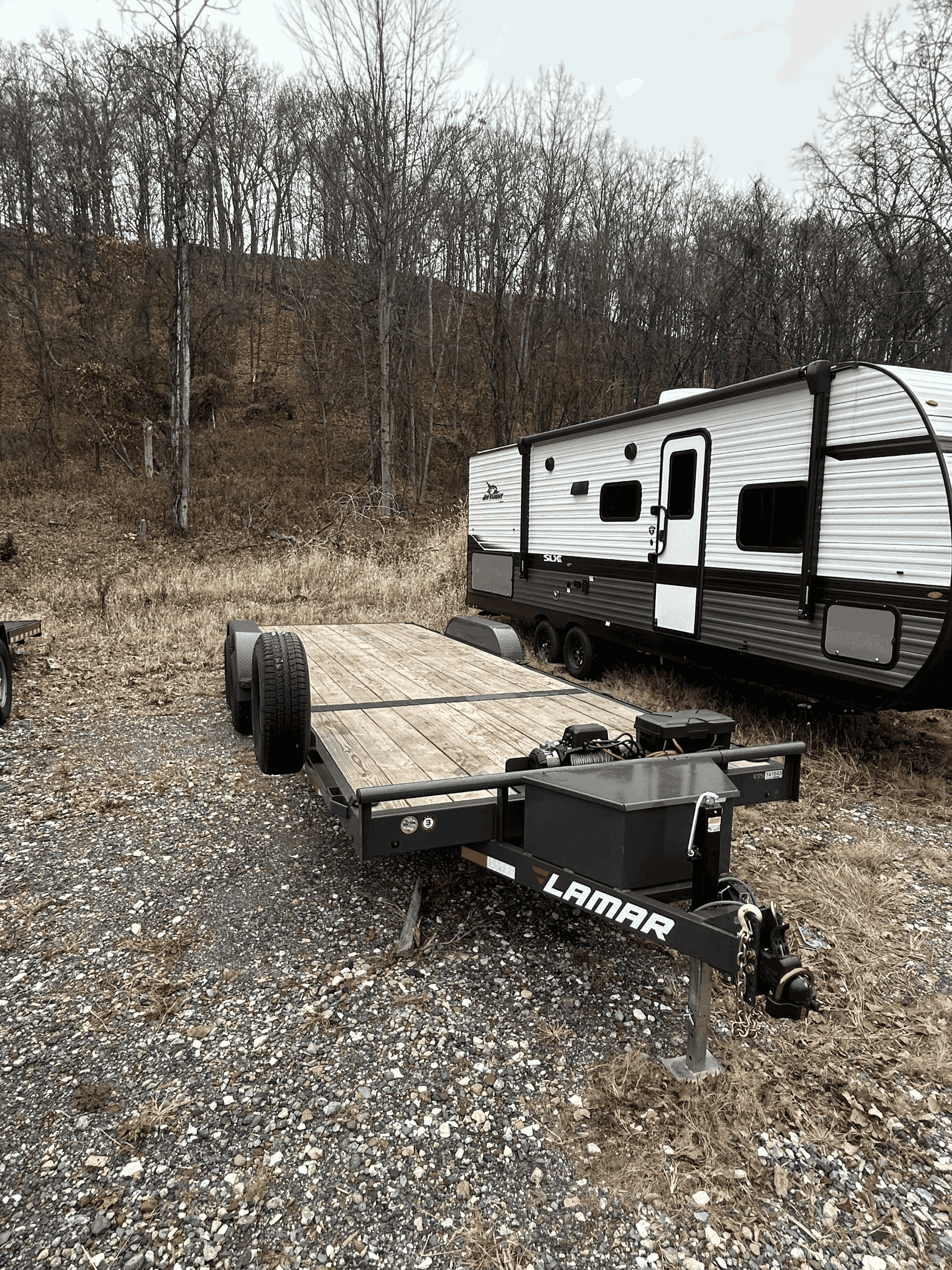 Empty flatbed trailer with a wood deck parked on a gravel surface in front of a white and black travel trailer, with a wooded hillside in the background.