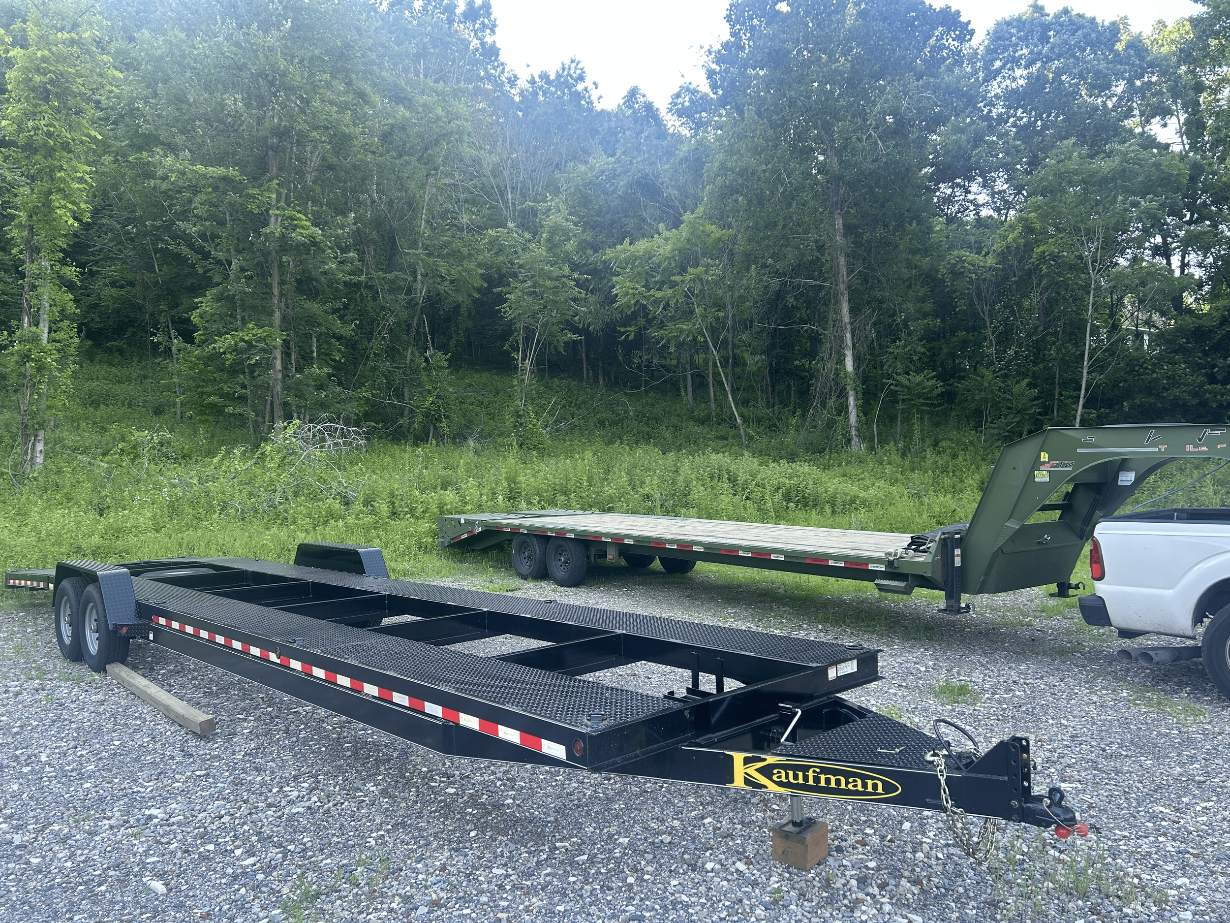 Two utility trailers parked on gravel ground in front of a wooded area. One trailer is black and empty, while the other is green with a ramp, attached to a white pickup truck.