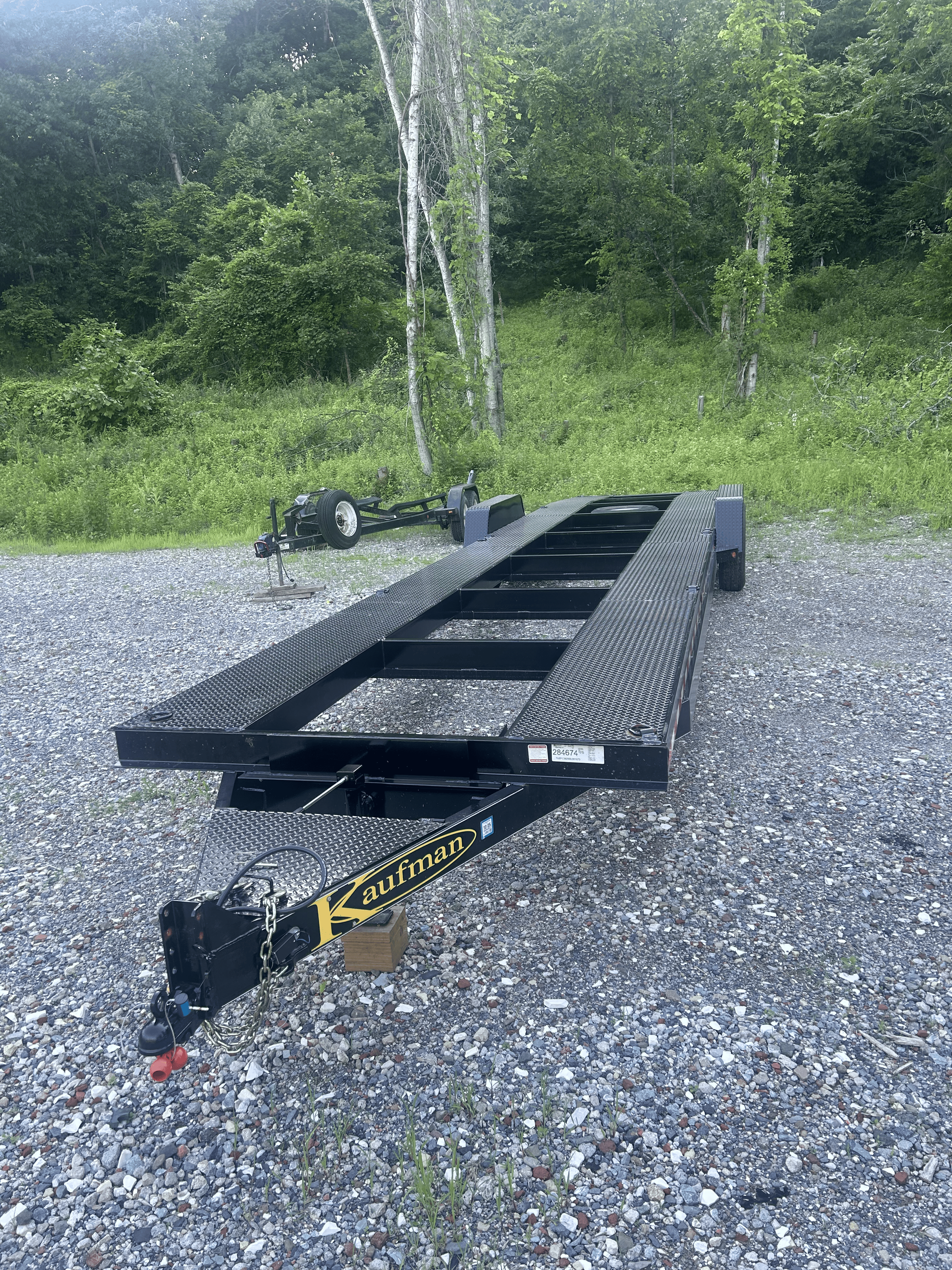 Black car hauler trailer on gravel ground with green trees and grass in the background.