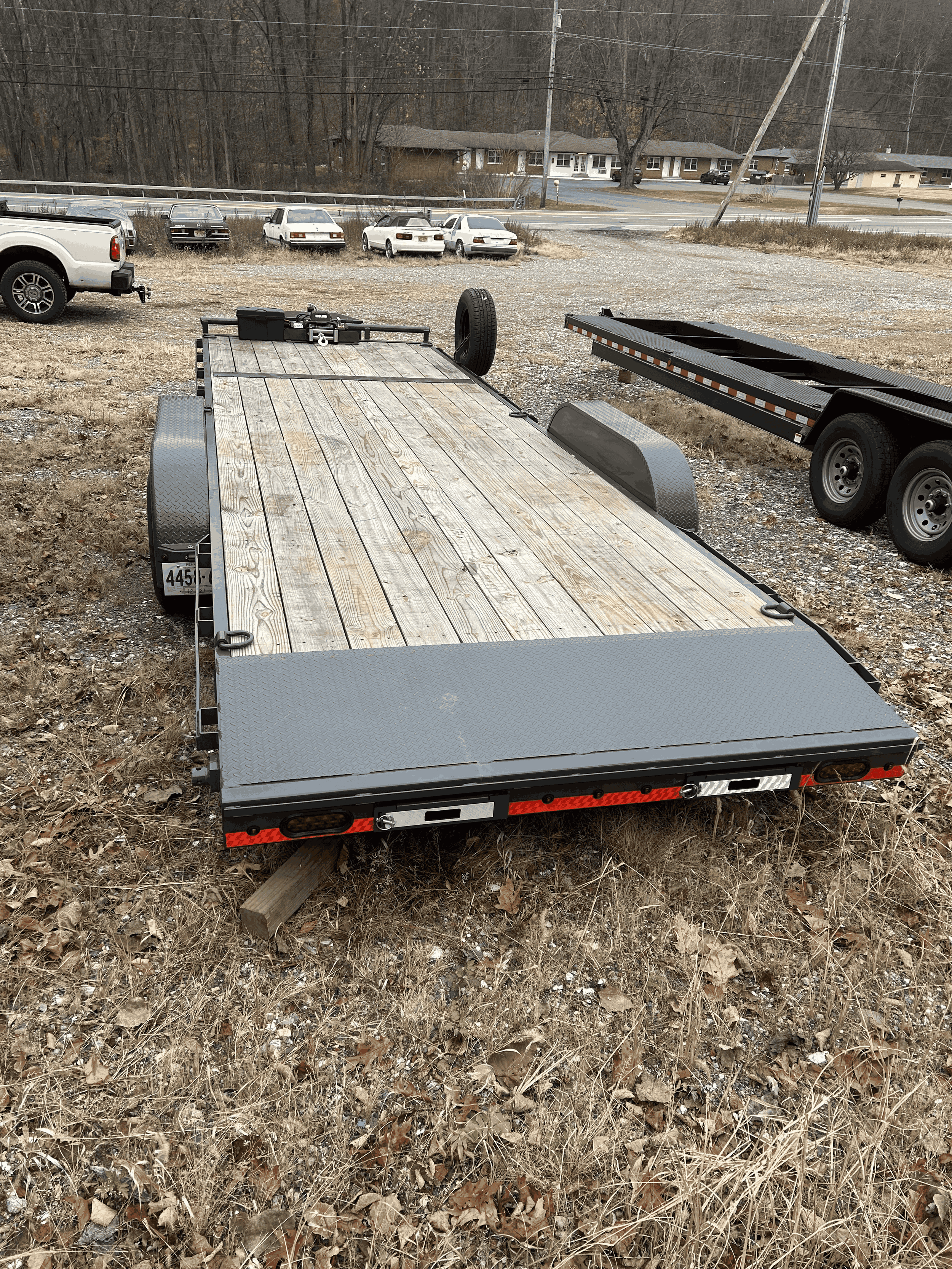 Empty flatbed trailer with a wooden deck parked on grassy, gravel area.