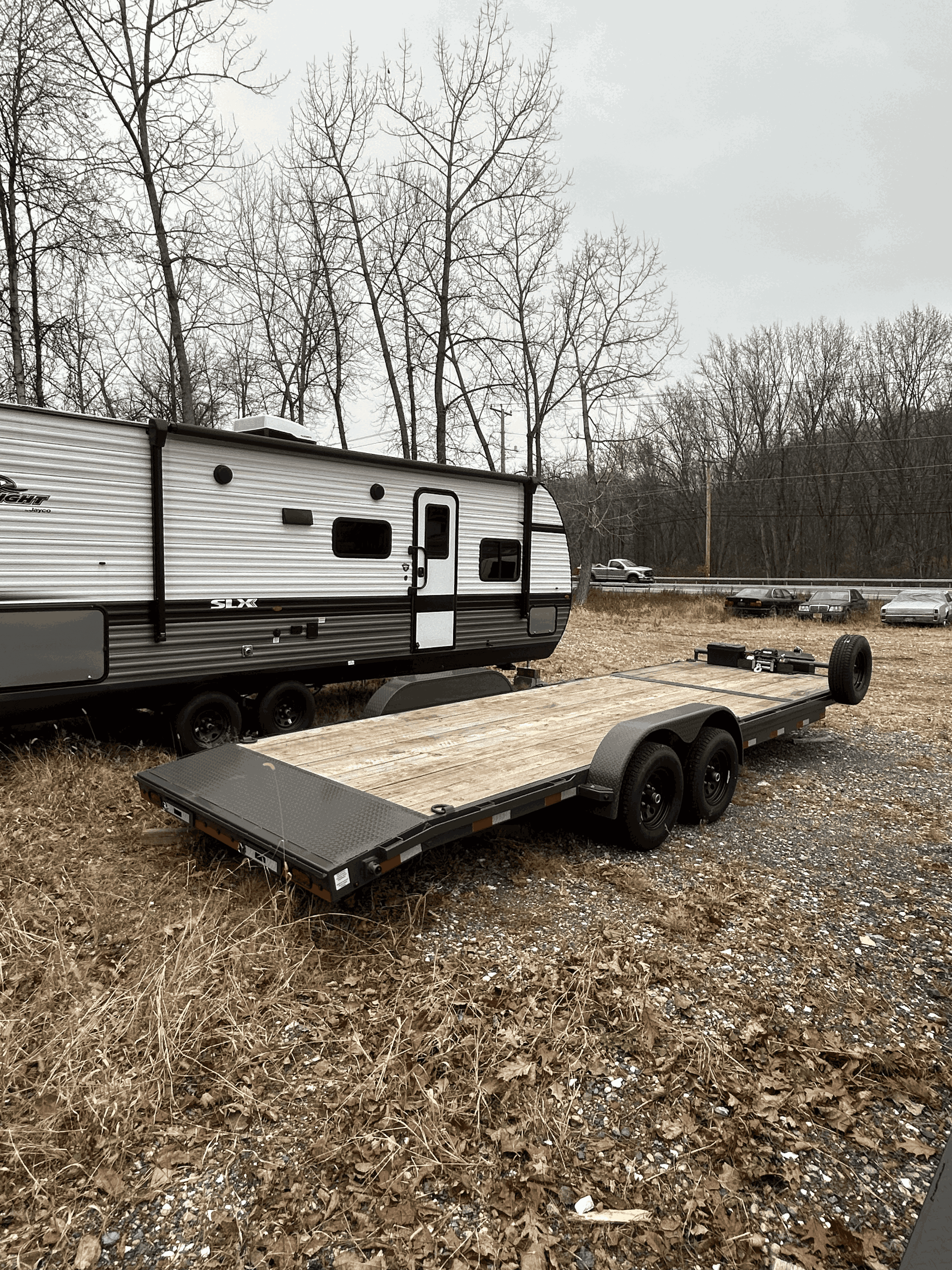 An empty flatbed trailer parked on a dirt and gravel lot, with a travel trailer and several cars parked in the background under a cloudy sky and leafless trees.