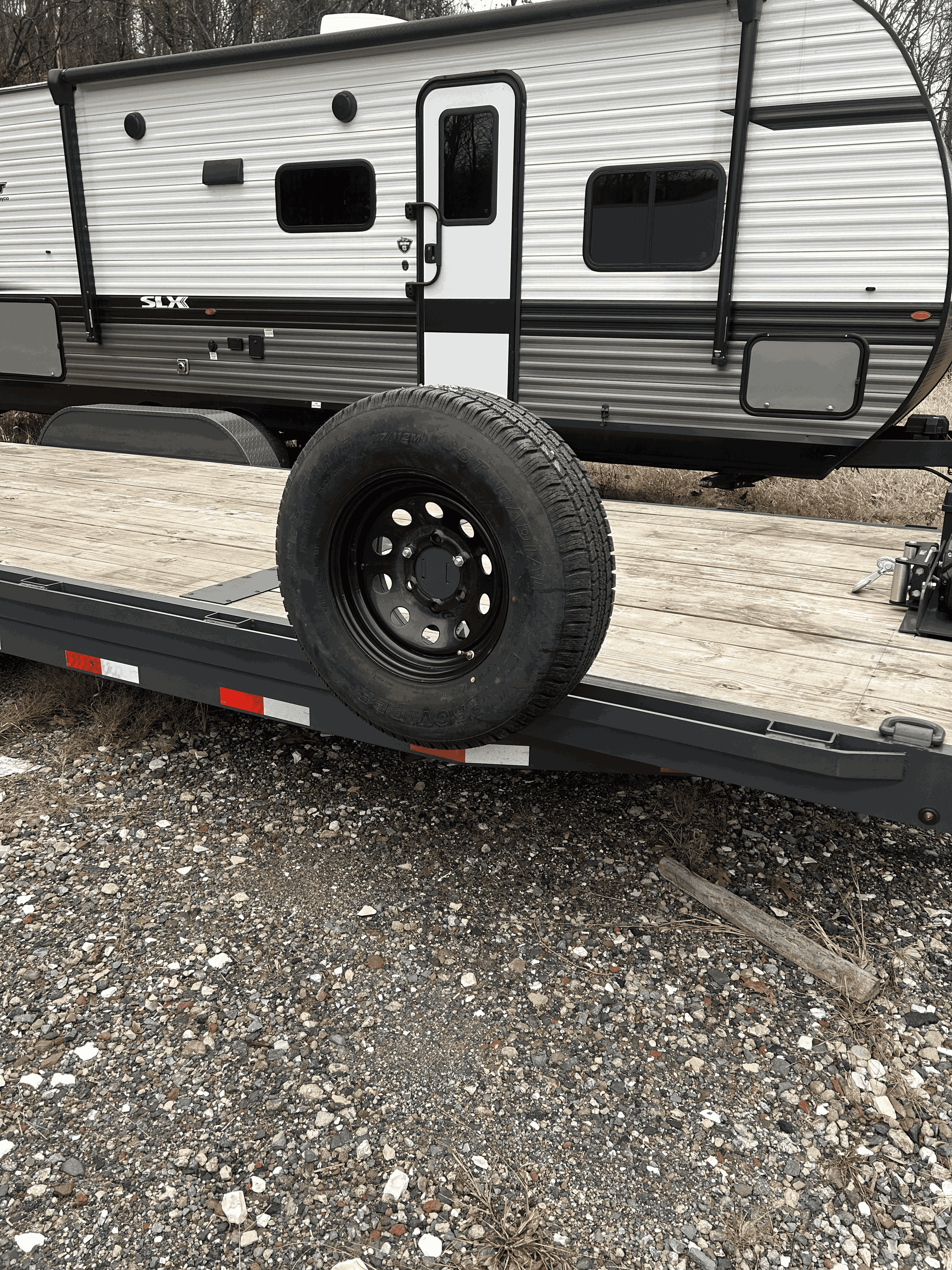 A black and white travel trailer on a flatbed trailer in an outdoor gravel area.