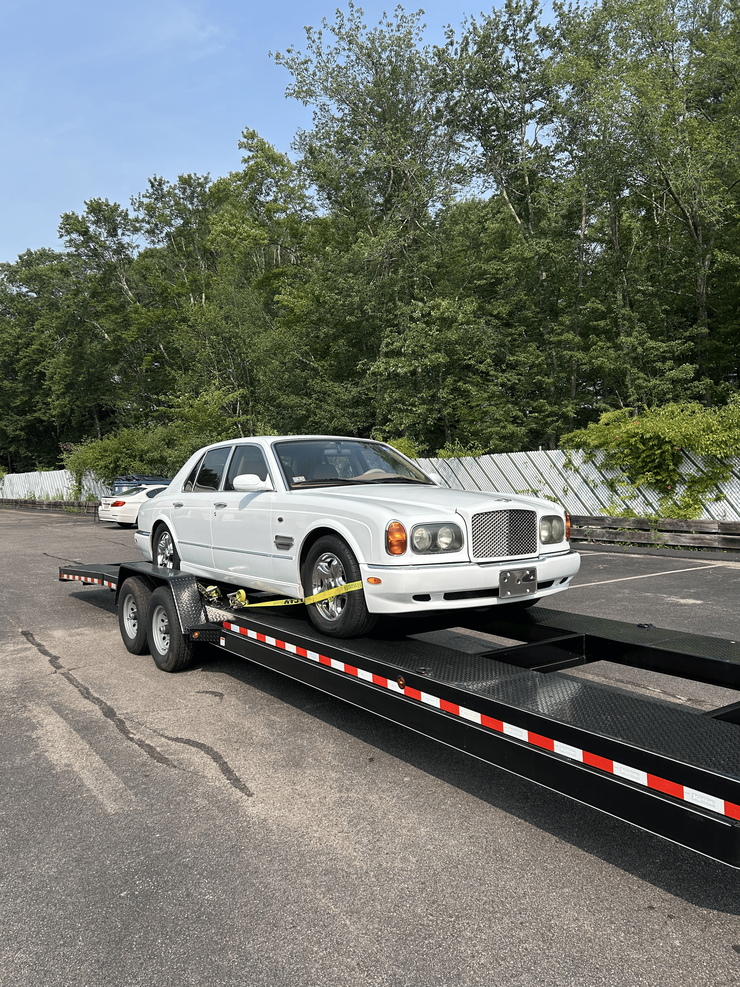 White vintage car on a flatbed trailer in an outdoor parking lot with trees in the background.