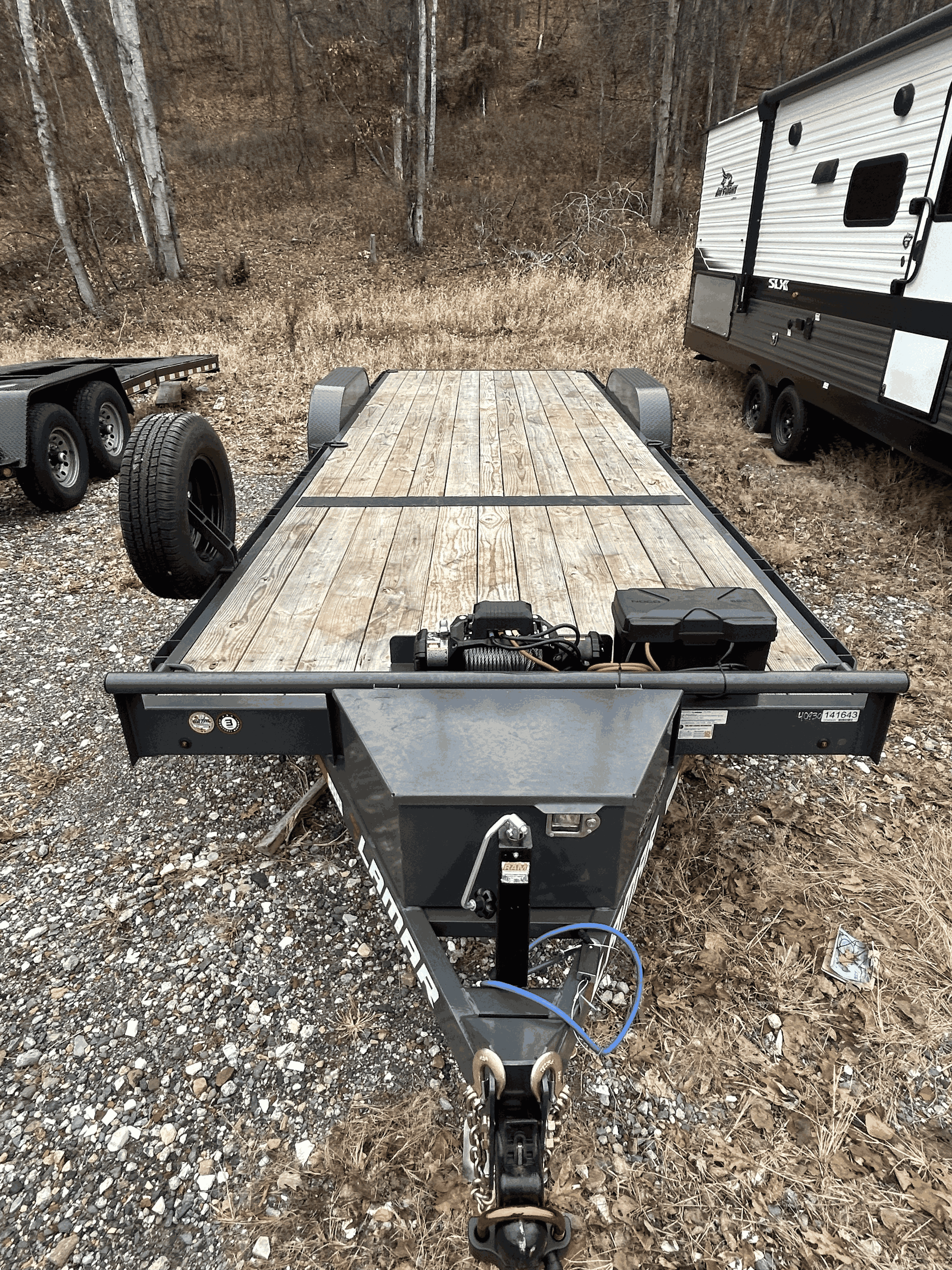 A flatbed utility trailer with a wooden deck, parked on a gravel area with leafless trees in the background.
