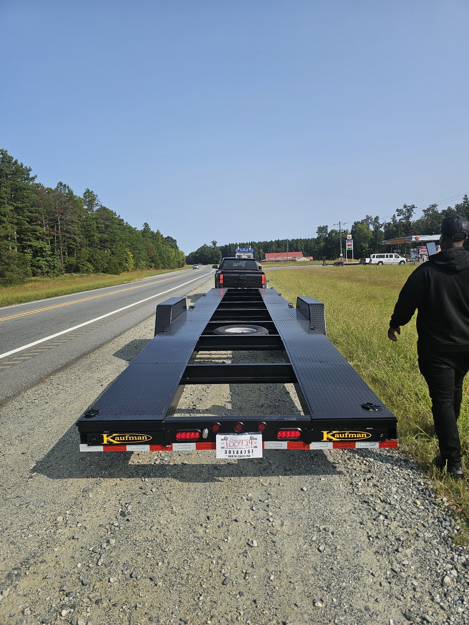 A black car being towed on a flatbed trailer along a rural road with trees on the side and a gas station in the background, under a clear blue sky.