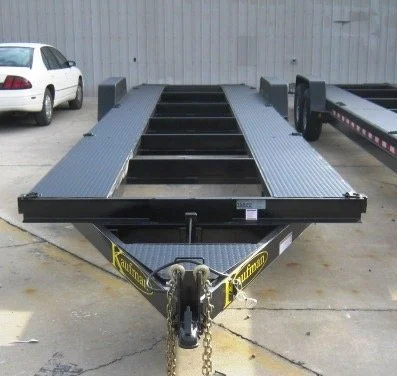 A black flatbed trailer with a ramp, parked on concrete, with a white SUV to the left and another trailer to the right, against a light gray metal wall