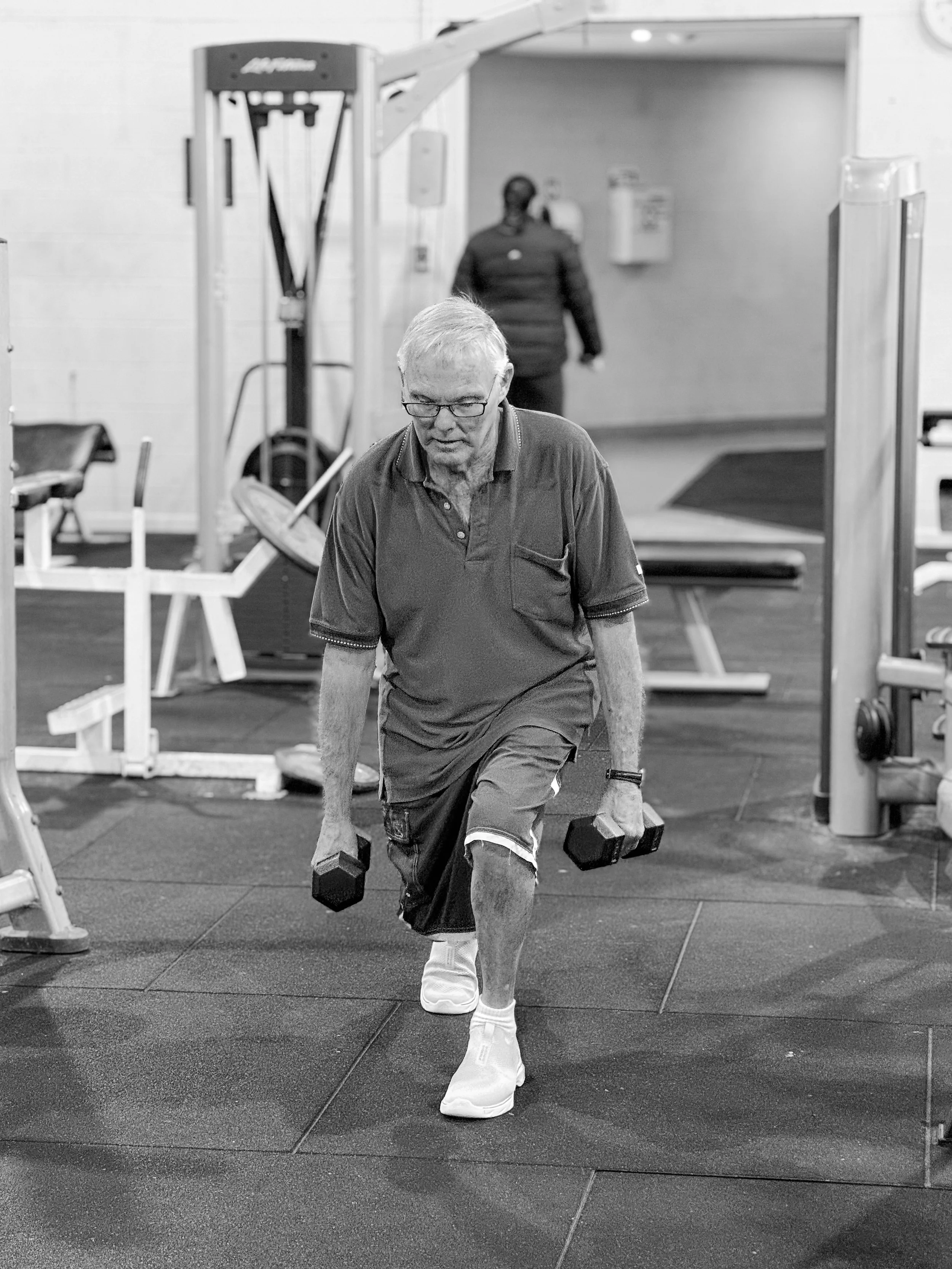 An elderly man working out in a gym, performing a lunge while holding dumbbells. He is wearing glasses, a polo shirt, and athletic shoes.