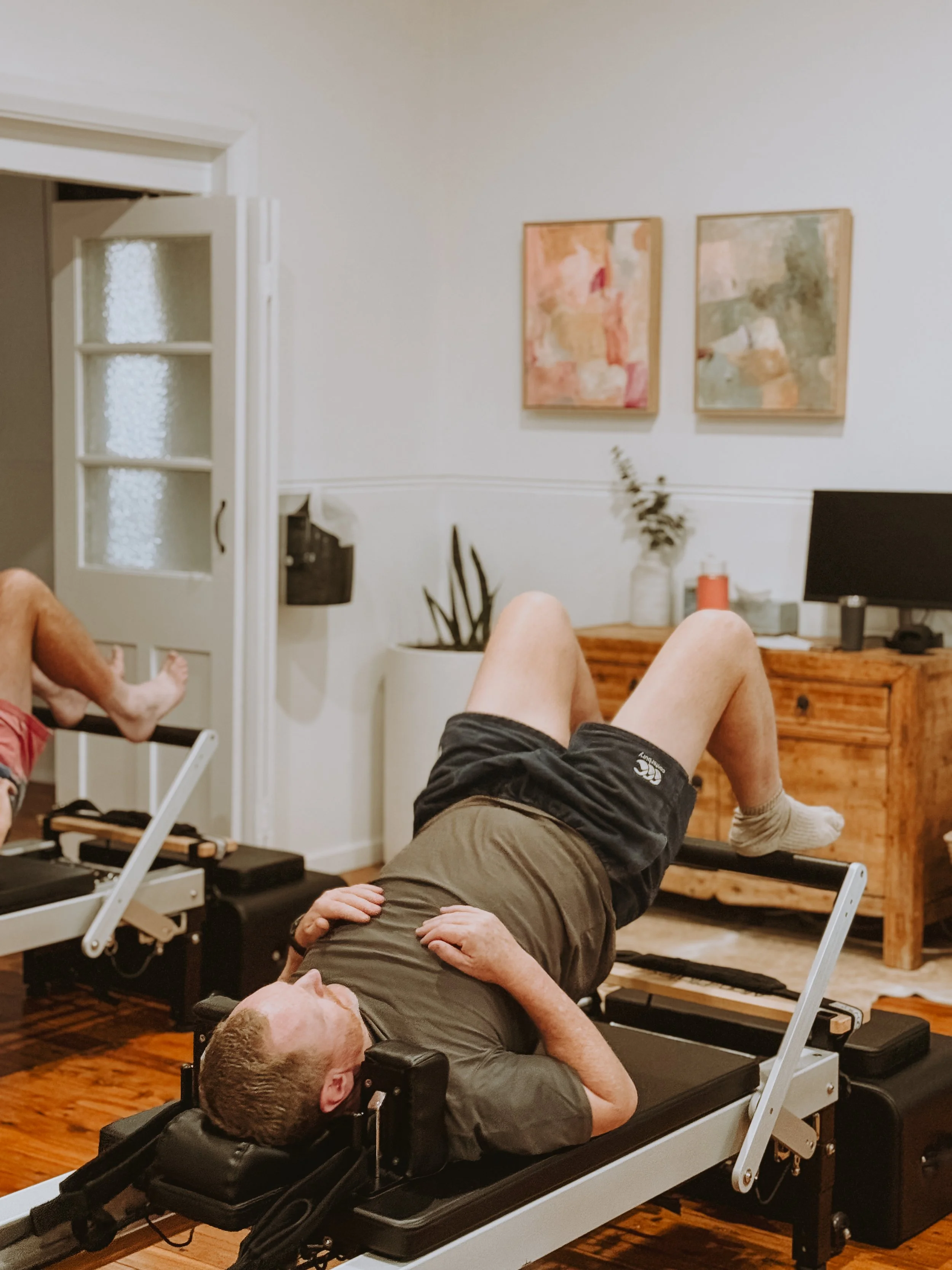 Person lying on a Pilates reformer machine in a living room with wall art, plants, and a TV in the background.