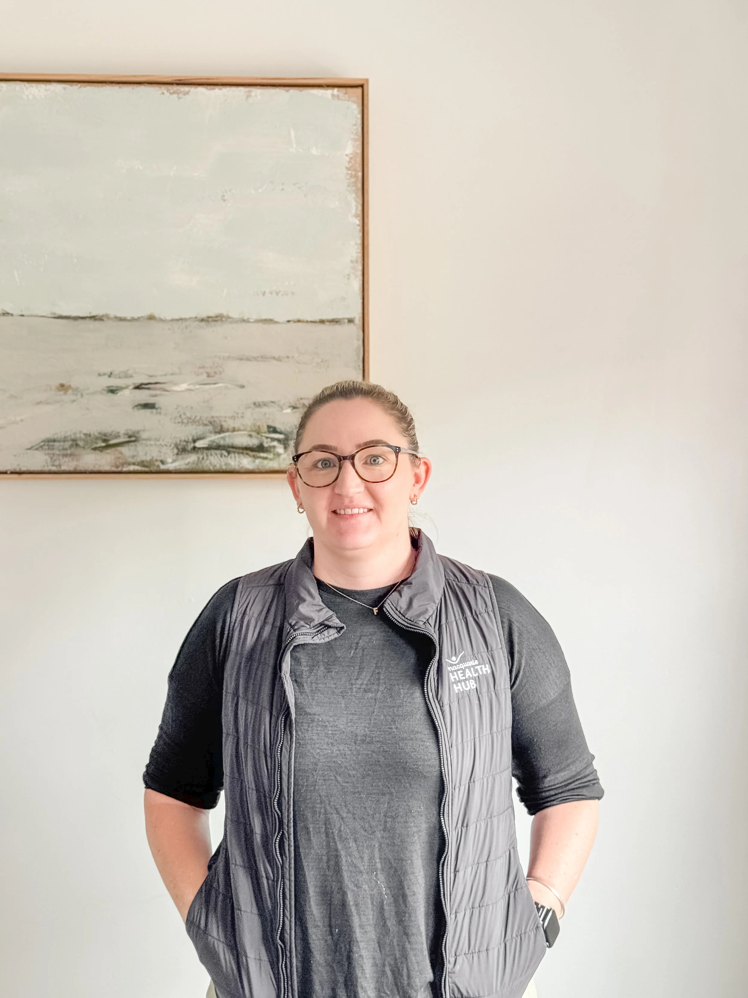 A woman standing indoors in front of a white wall with a framed abstract painting. She is wearing glasses, a dark gray shirt, and a black puffer vest with the logo 'Health Hub.' She has her hair tied back and is smiling at the camera.
