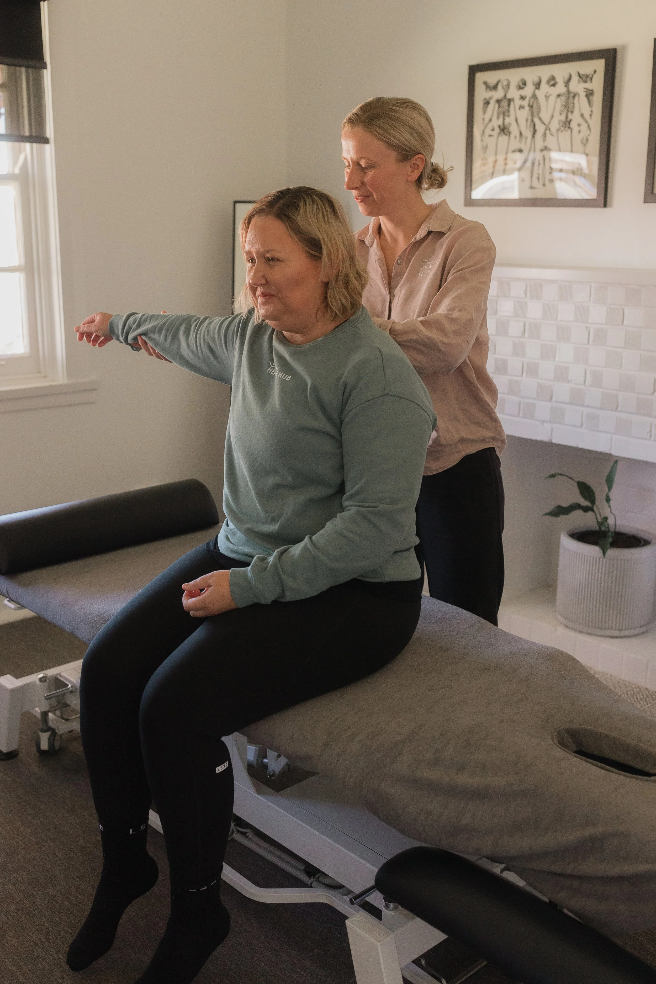 A woman sitting on a therapy table with her arm extended forward, assisted by a healthcare professional standing behind her, in a clinical setting.