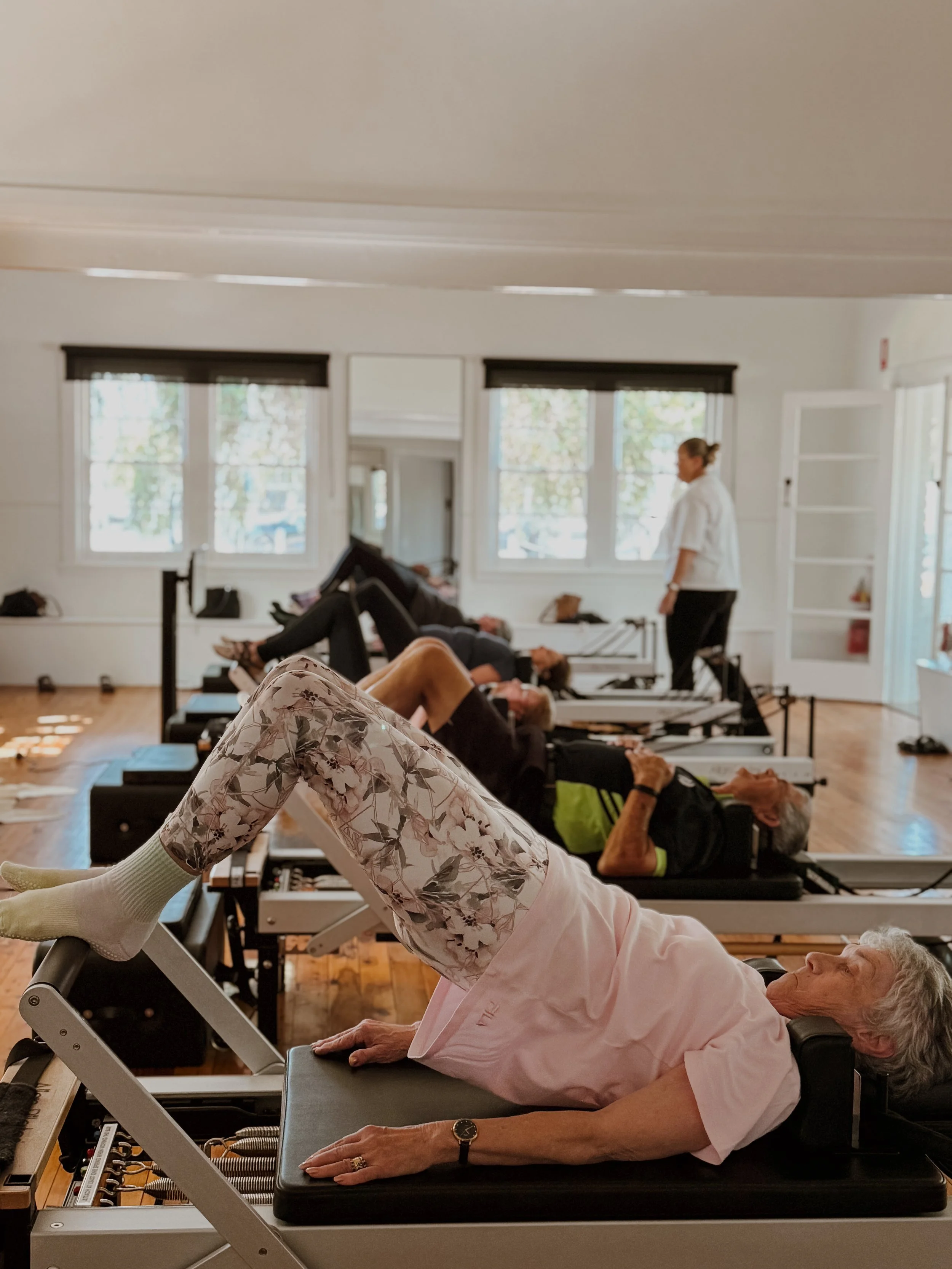 Older woman lying on a Pilates reformer machine in a fitness class, with other people and an instructor in the background.