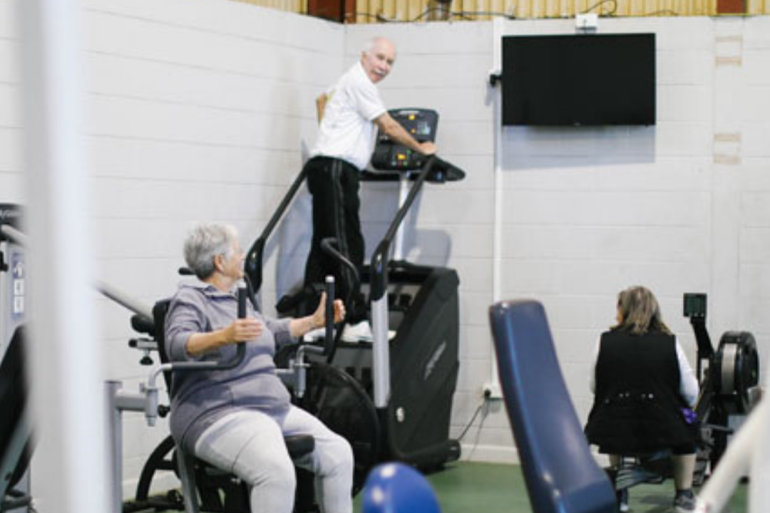 Three elderly individuals in a gym using exercise equipment, with a man walking on a treadmill, a woman on a seated exercise machine, and another person on a stationary bike.