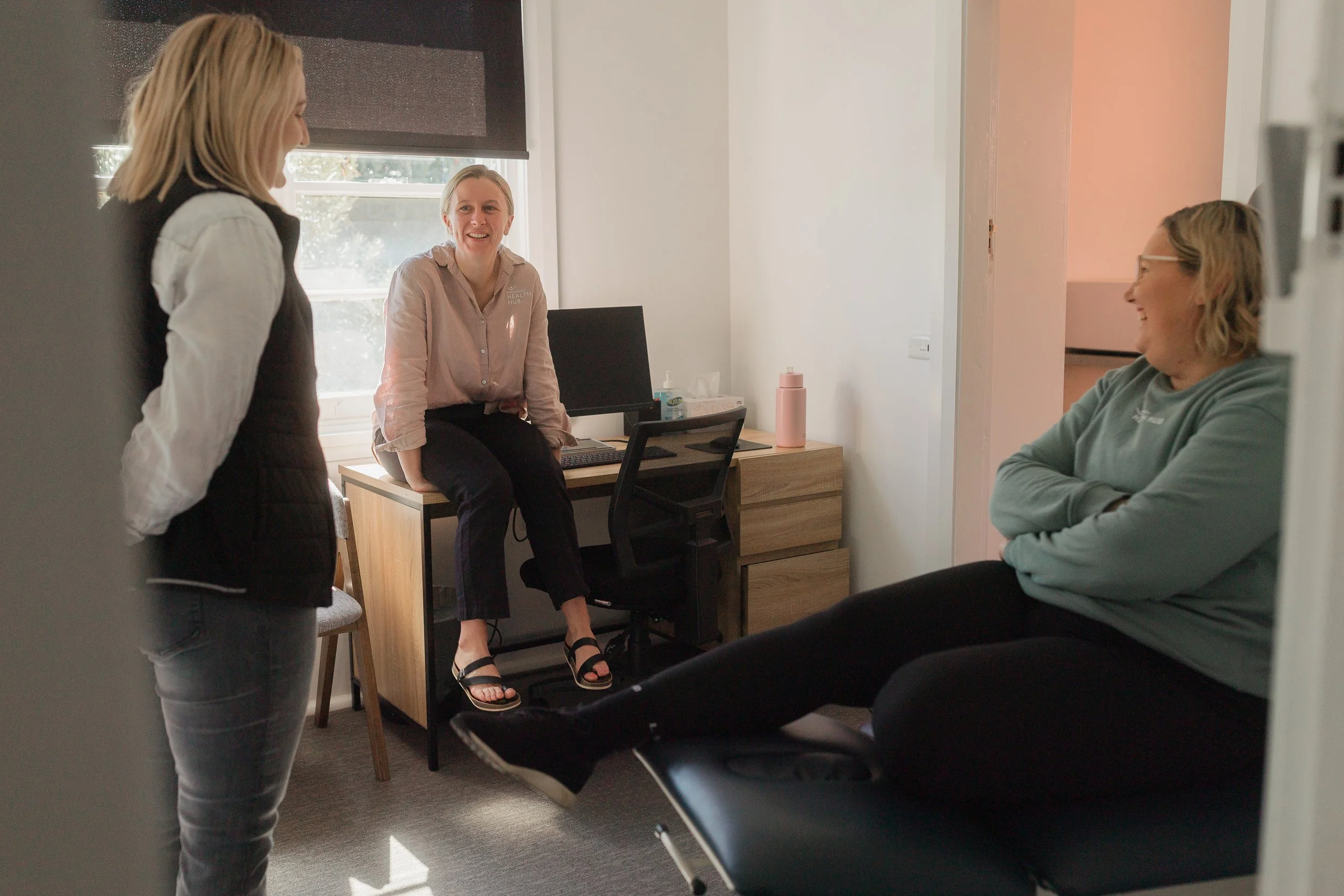 Three women in an office, smiling and engaging in a friendly conversation. One woman is seated on a desk, another is sitting on a chair, and the third woman stands with her back partially to the camera.