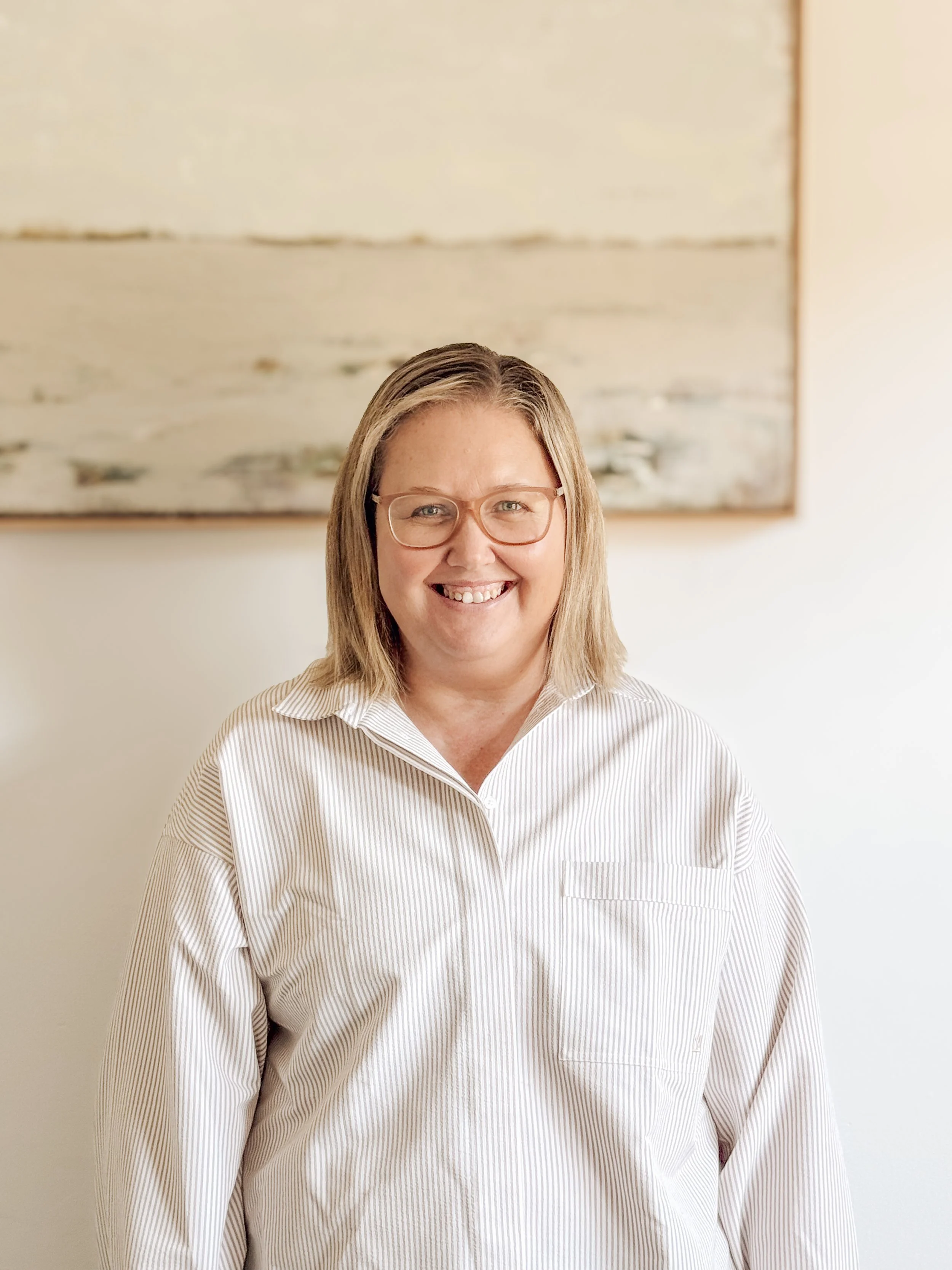 A woman with shoulder-length blonde hair wearing glasses and a white striped button-up shirt smiling in front of a light-colored wall and abstract artwork.