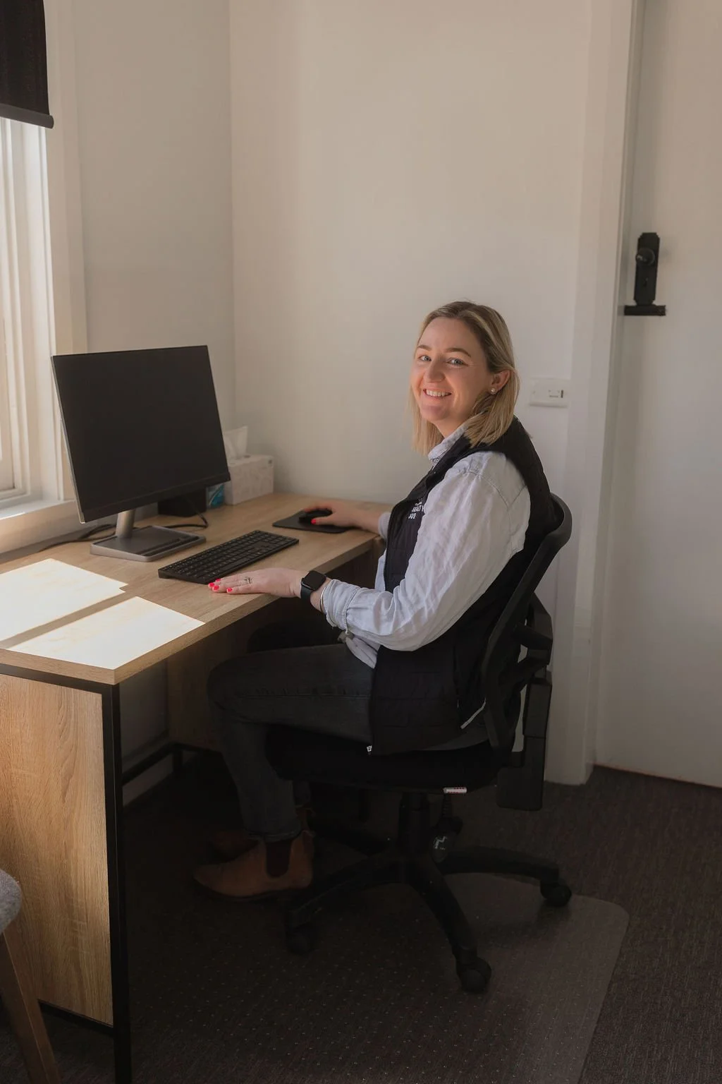 A woman sitting at a desk with a computer in a well-lit office, smiling and looking at the camera.