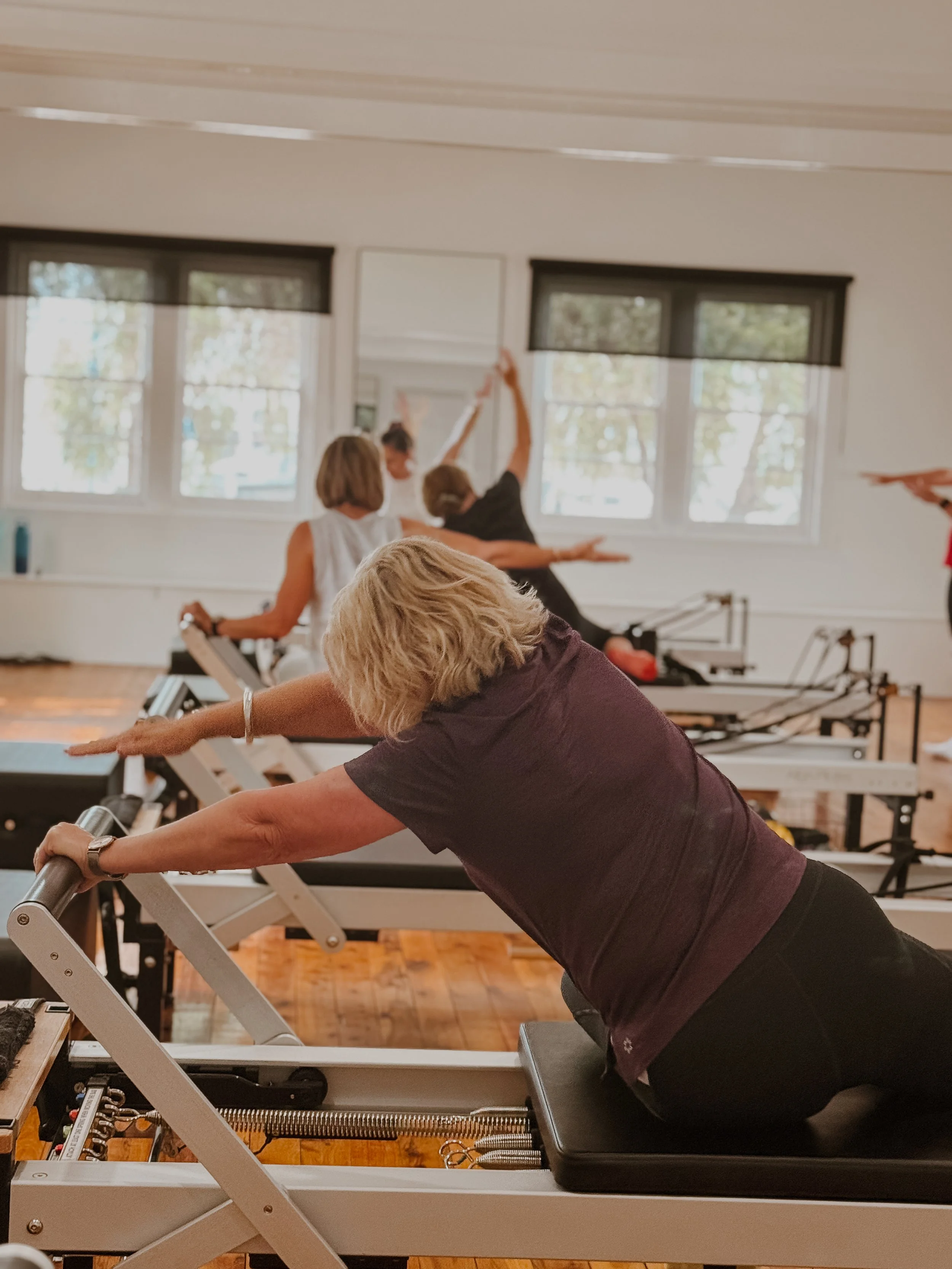 A group of women participating in a Pilates class using reformer machines in a well-lit studio with large windows.
