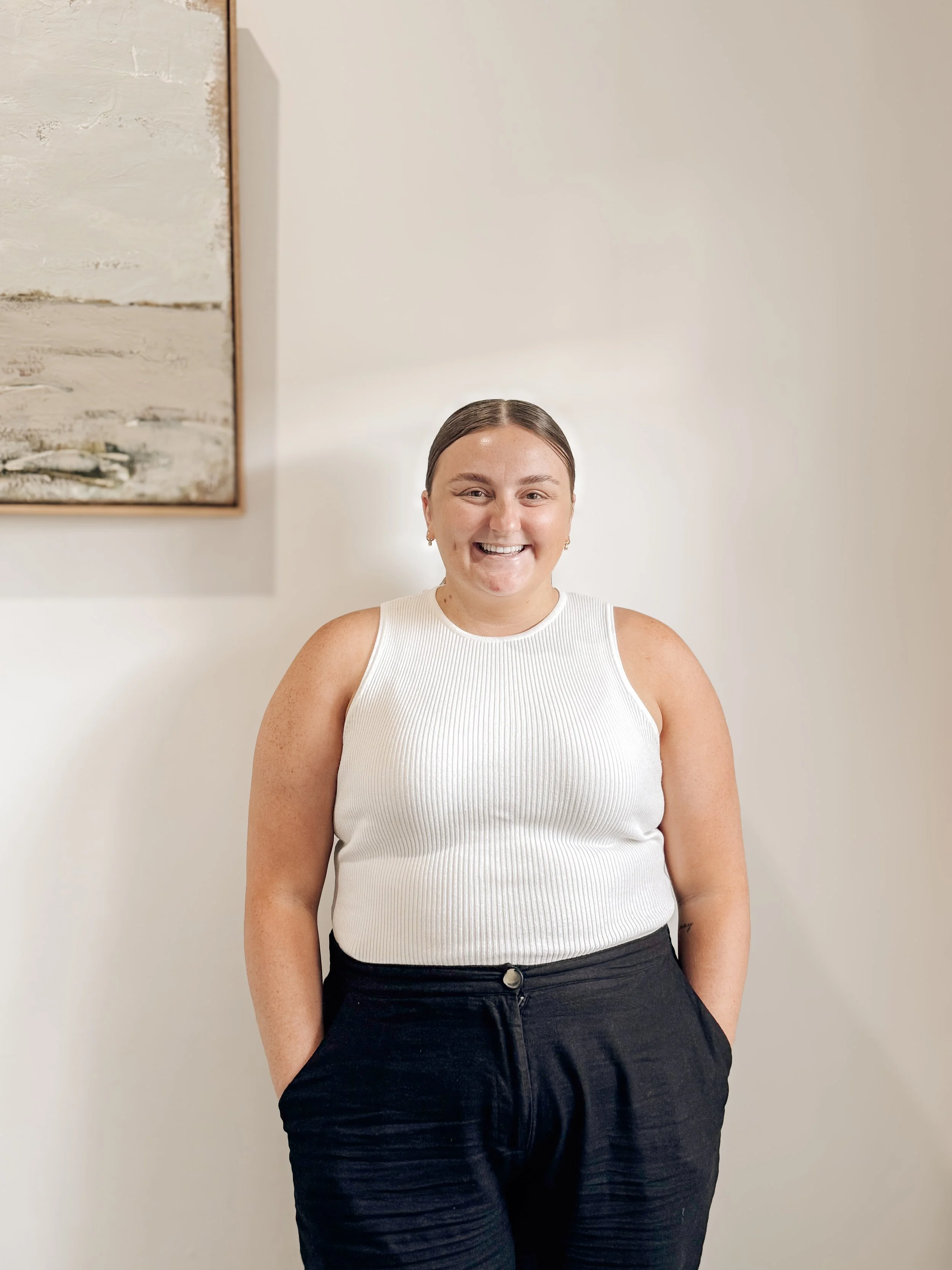 A woman smiling with her hands in her pockets, standing in front of a plain wall with a partially visible painting on the left.