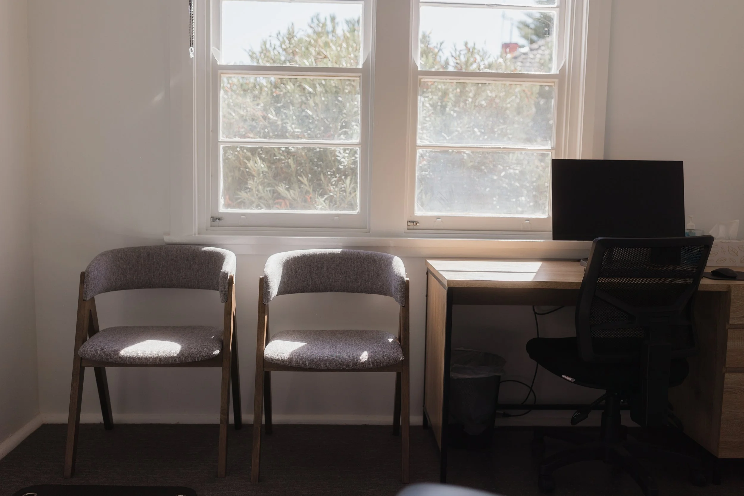 An office with two gray upholstered chairs, a desk with a computer monitor, a black office chair, a window showing a view of trees, and sunlight casting shadows on the chairs and desk.