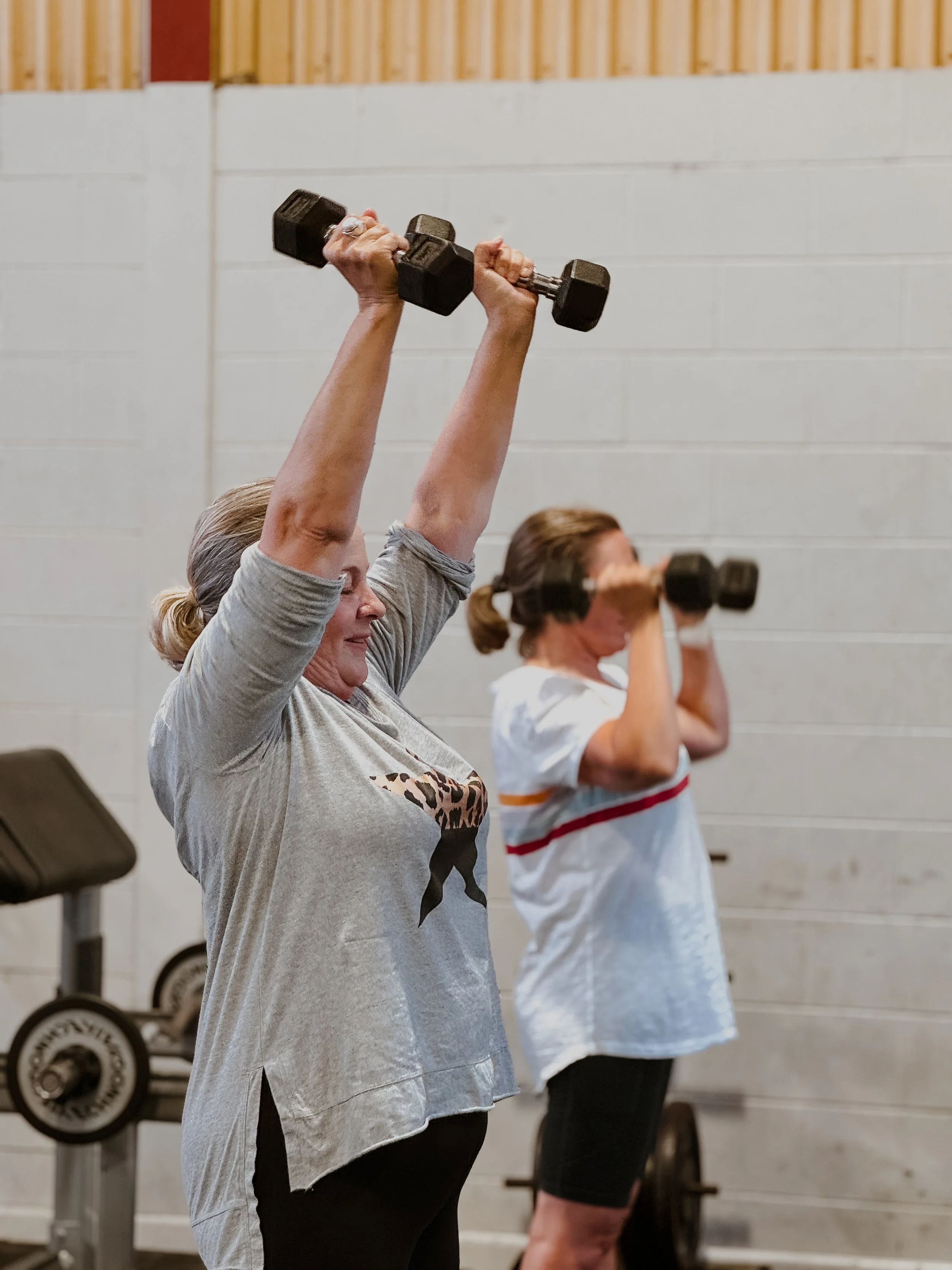 Two women lifting dumbbells overhead during a workout in a gym.