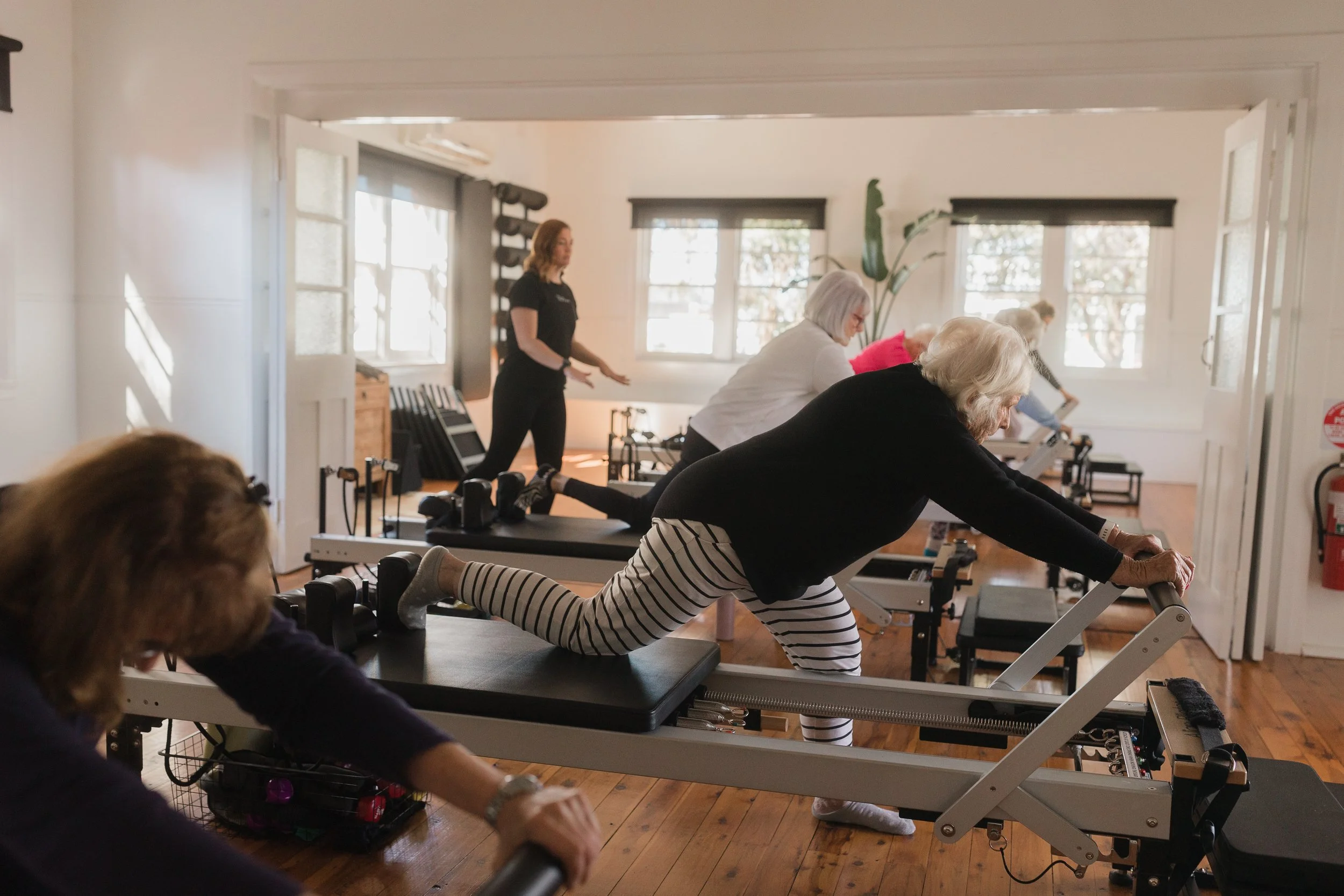 Older women doing Pilates exercises on reformer machines in a fitness class, instructor leading in the background.