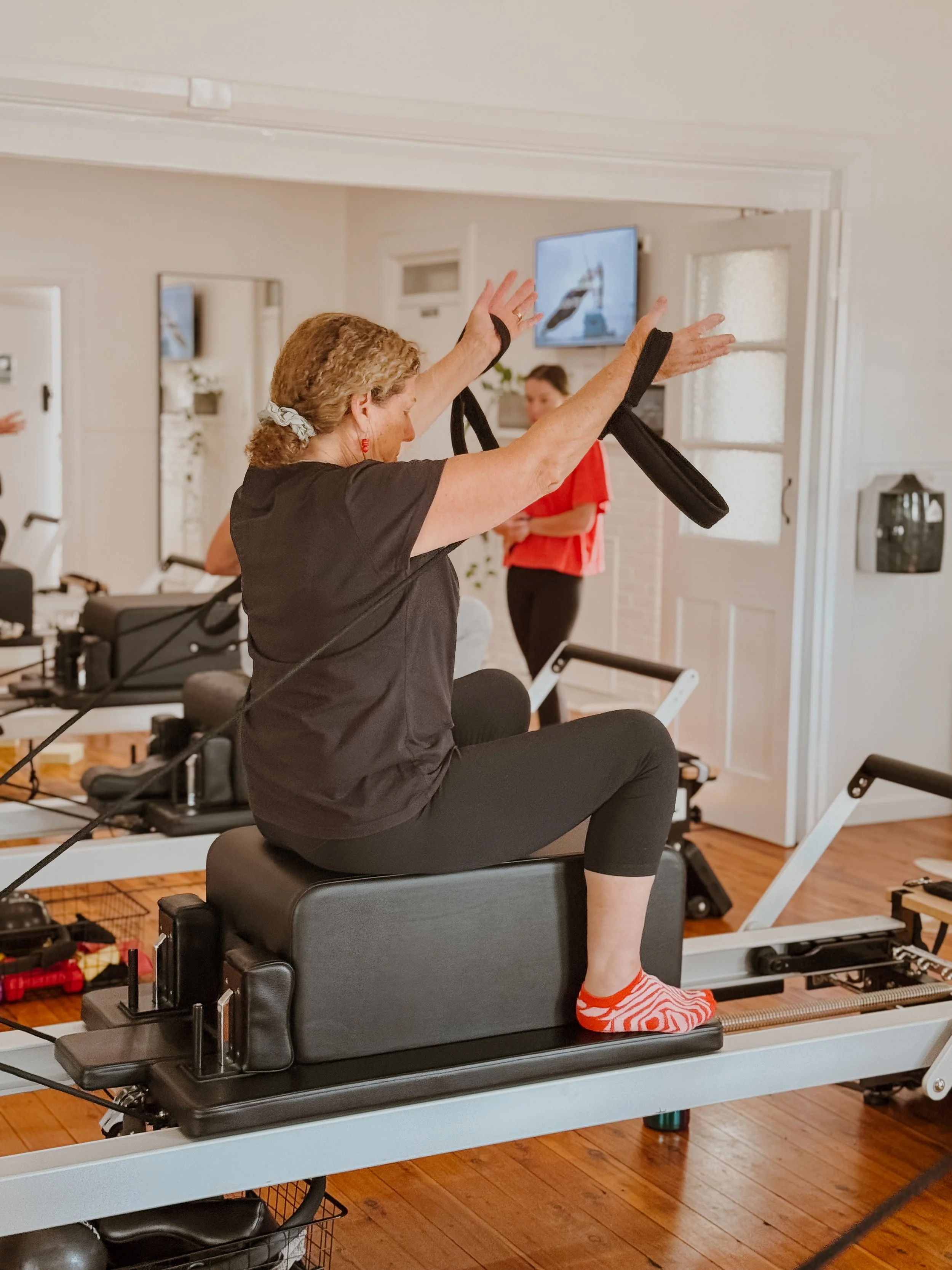 Woman exercising on a Pilates reformer machine, seated with arms raised, in a home gym setting.