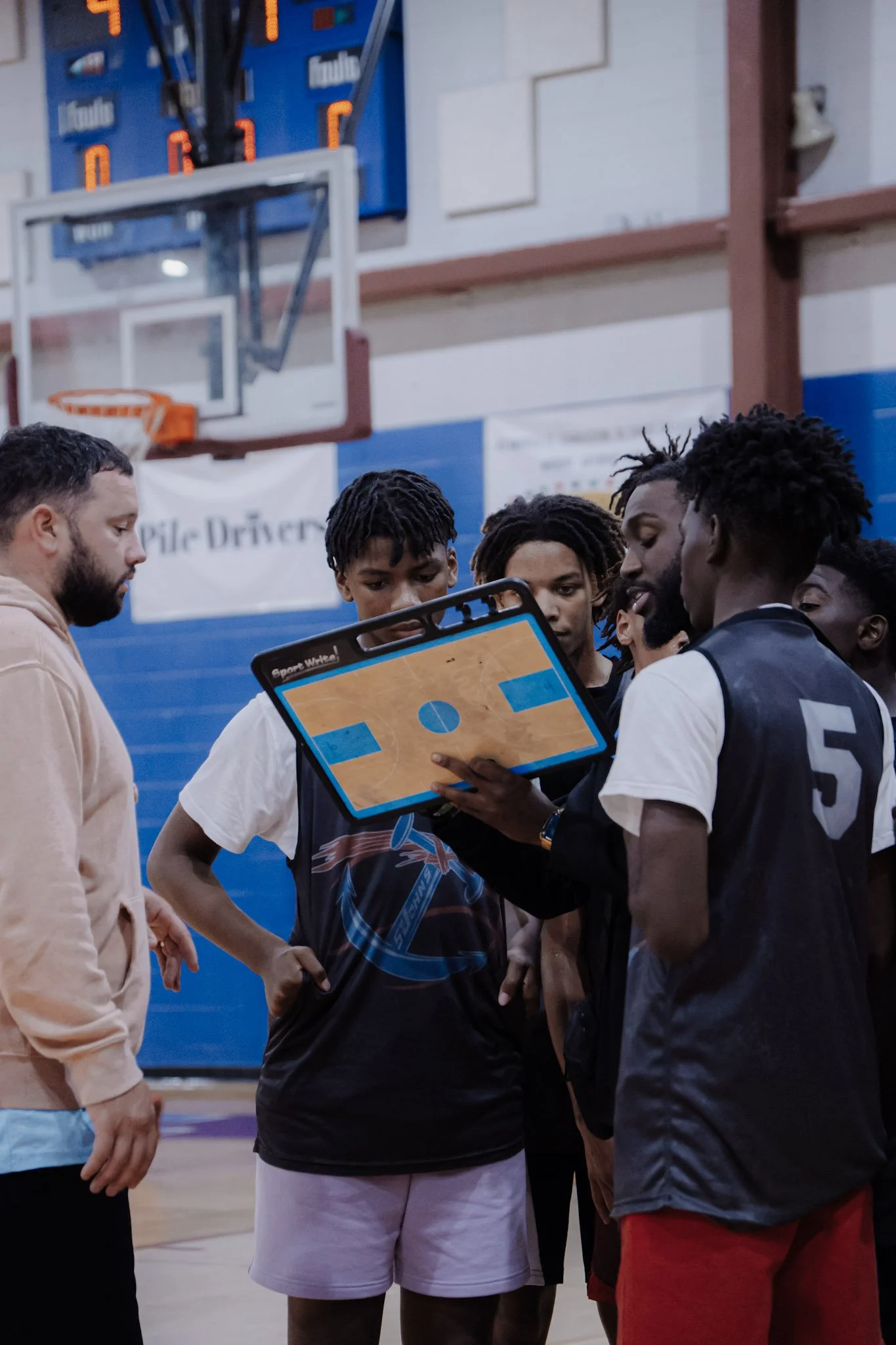 Basketball team huddling during a timeout, with coach showing play diagram on a tablet, inside gymnasium with basketball hoop in background.