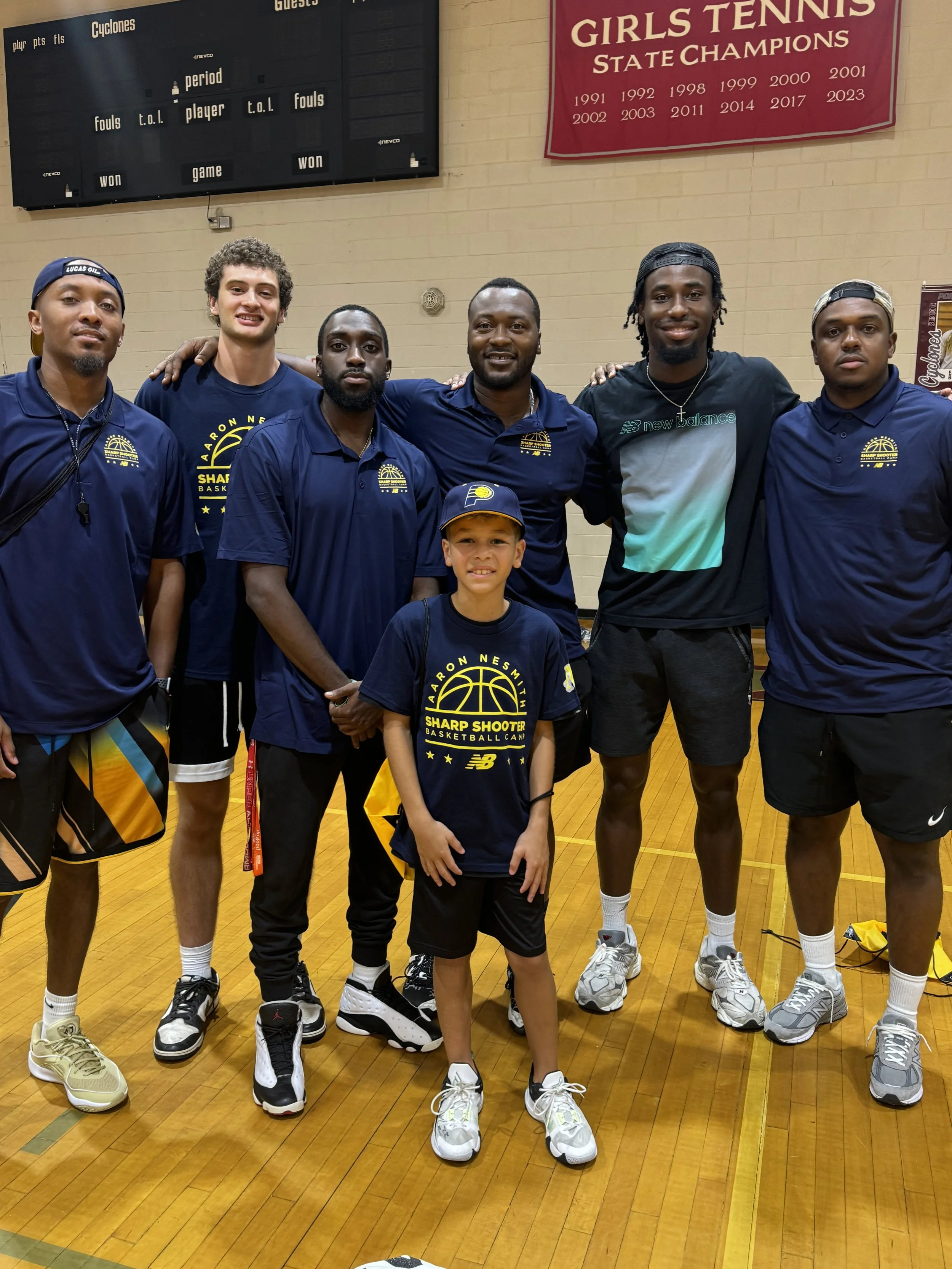 Group of five basketball players and one young boy standing on a basketball court, wearing Sports apparel with logos, smiling for a photo. Banner in the background reads "Girls Tennis State Champions."