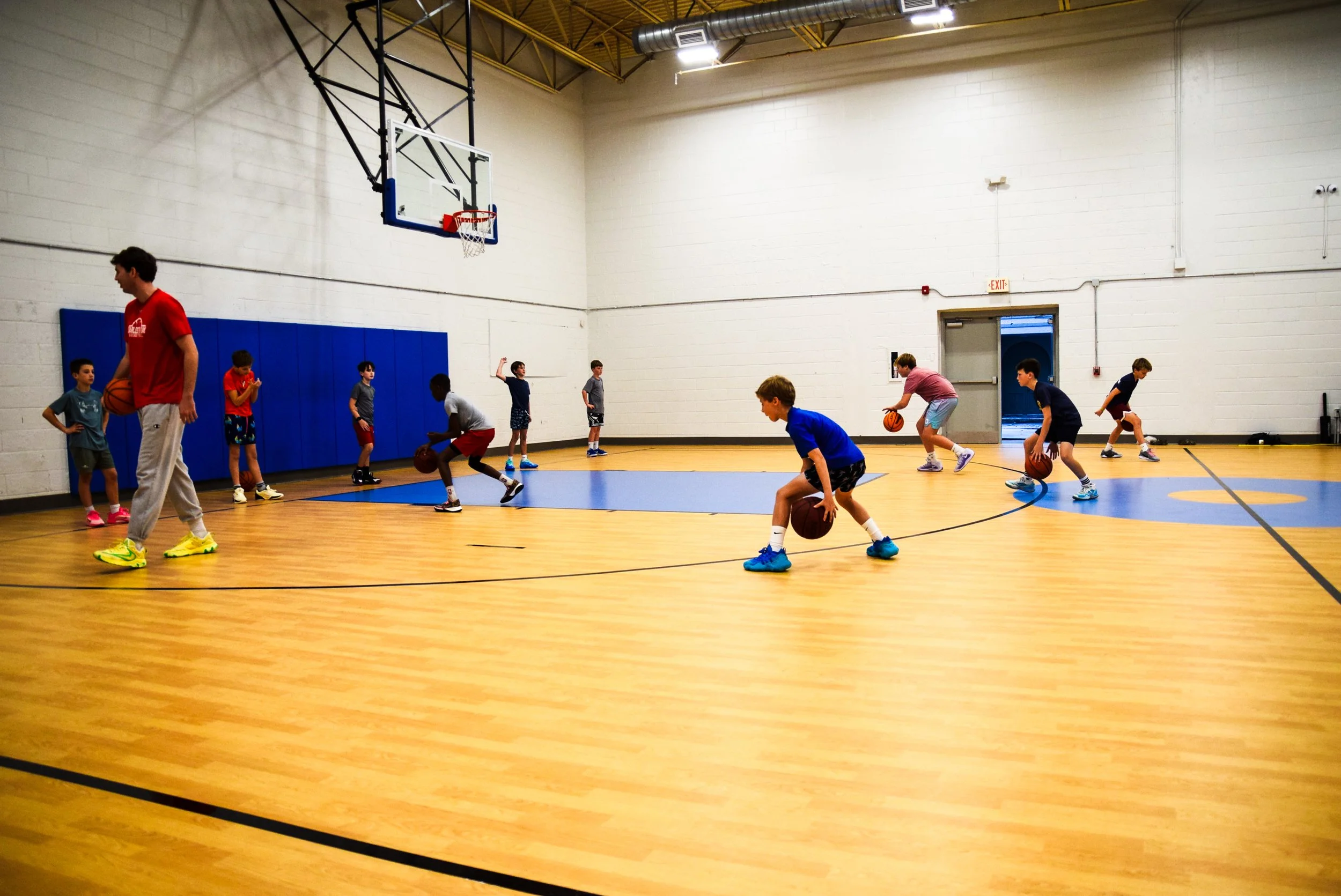 Young boys practicing basketball drills in an indoor gym, with a coach supervising.