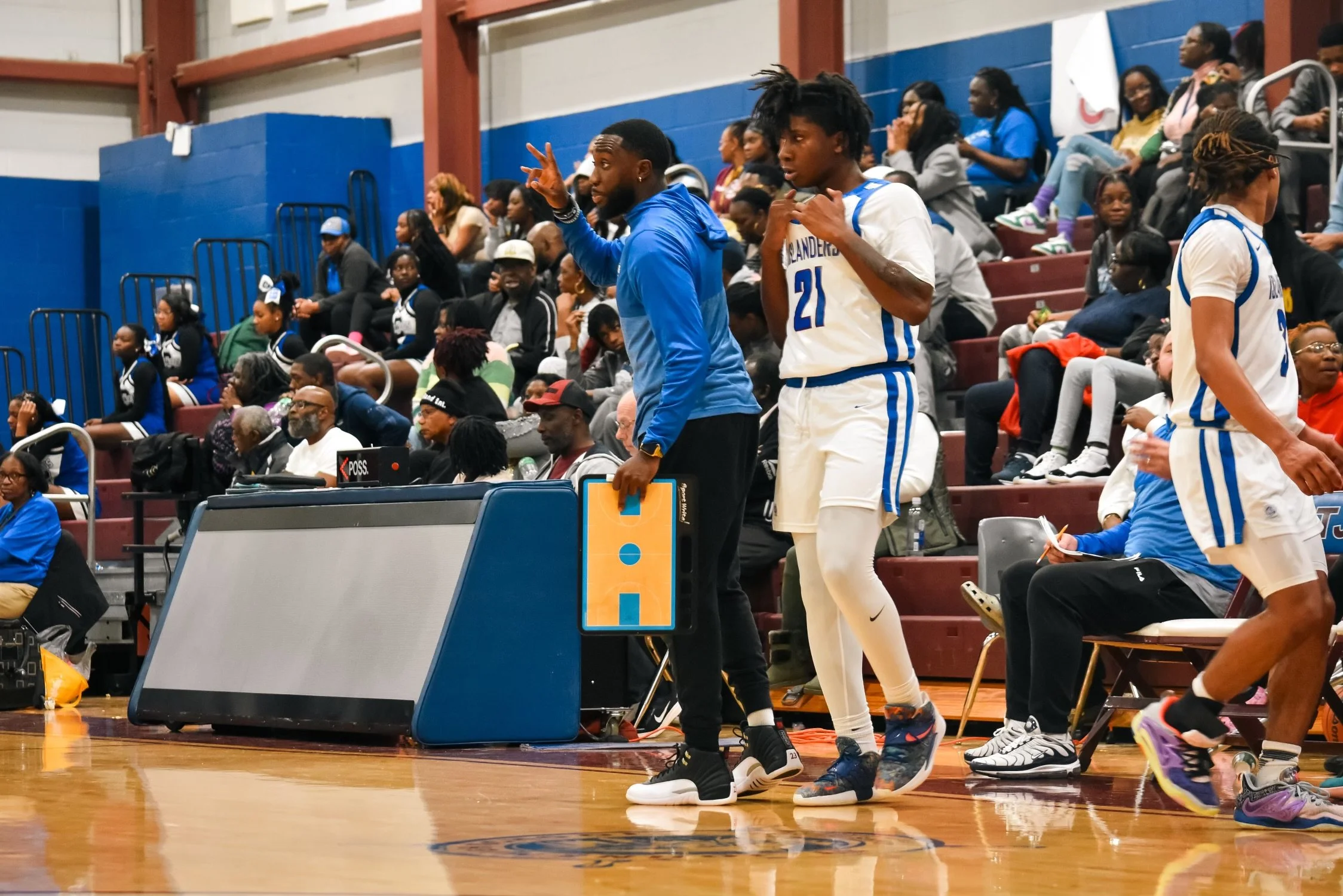 Basketball players and coaches on the sideline during a game, with spectators seated in the background.