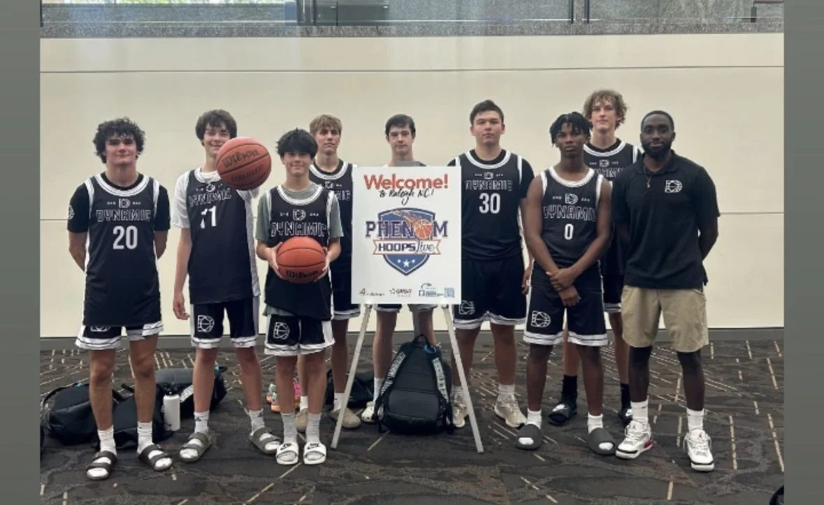 A youth basketball team in black uniforms standing for a photo indoors, with a coach on the right, holding two basketballs and standing behind a sign that reads 'Welcome! PHENOM HOOPS LIVE.' The team members are wearing matching uniforms with numbers