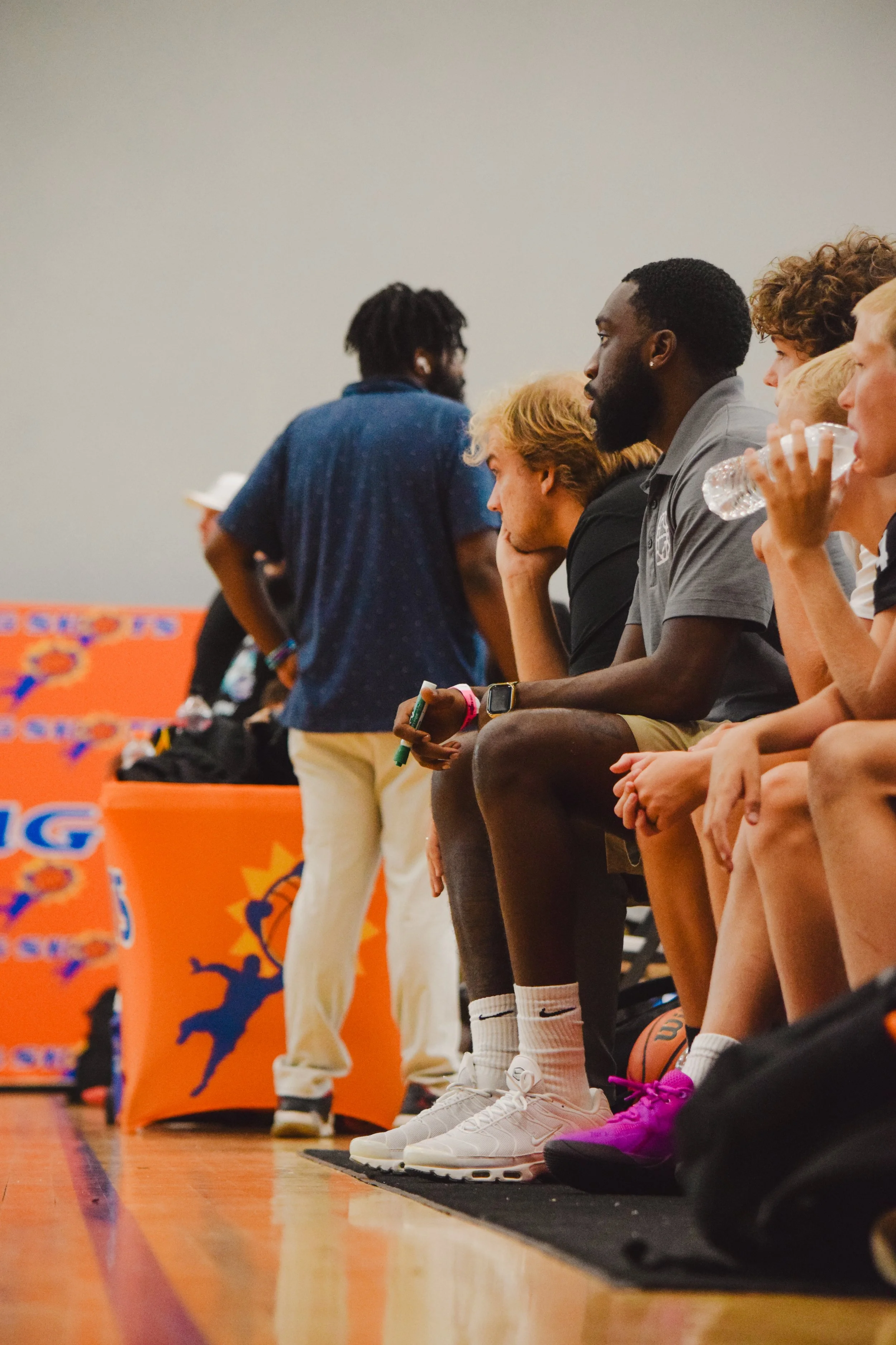 A group of young men sitting on a bench, some holding water bottles, at an indoor sports event with orange banners featuring a basketball logo in the background.