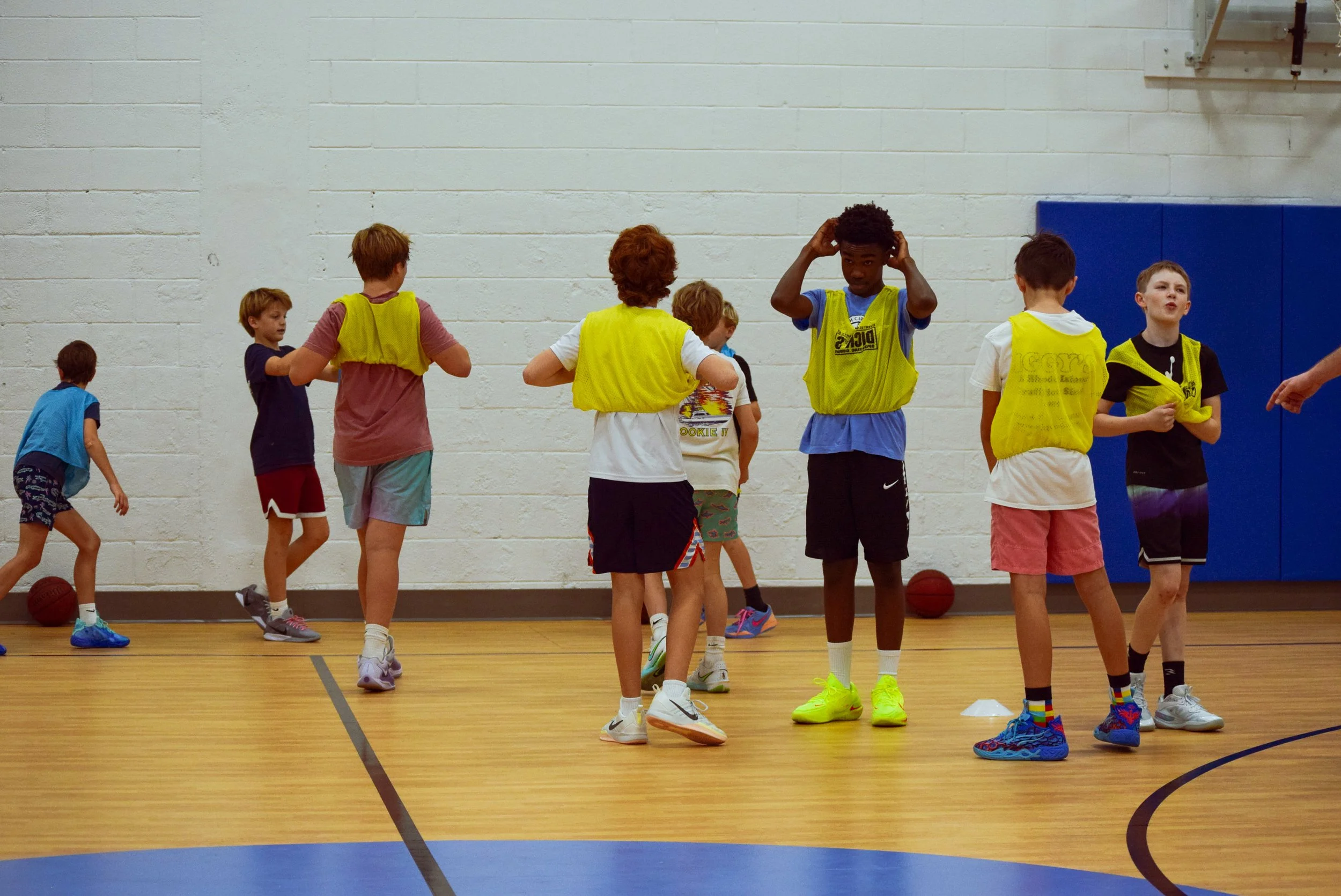 Children participating in a basketball practice in gym, some wearing yellow pinnies, with basketballs on the floor.