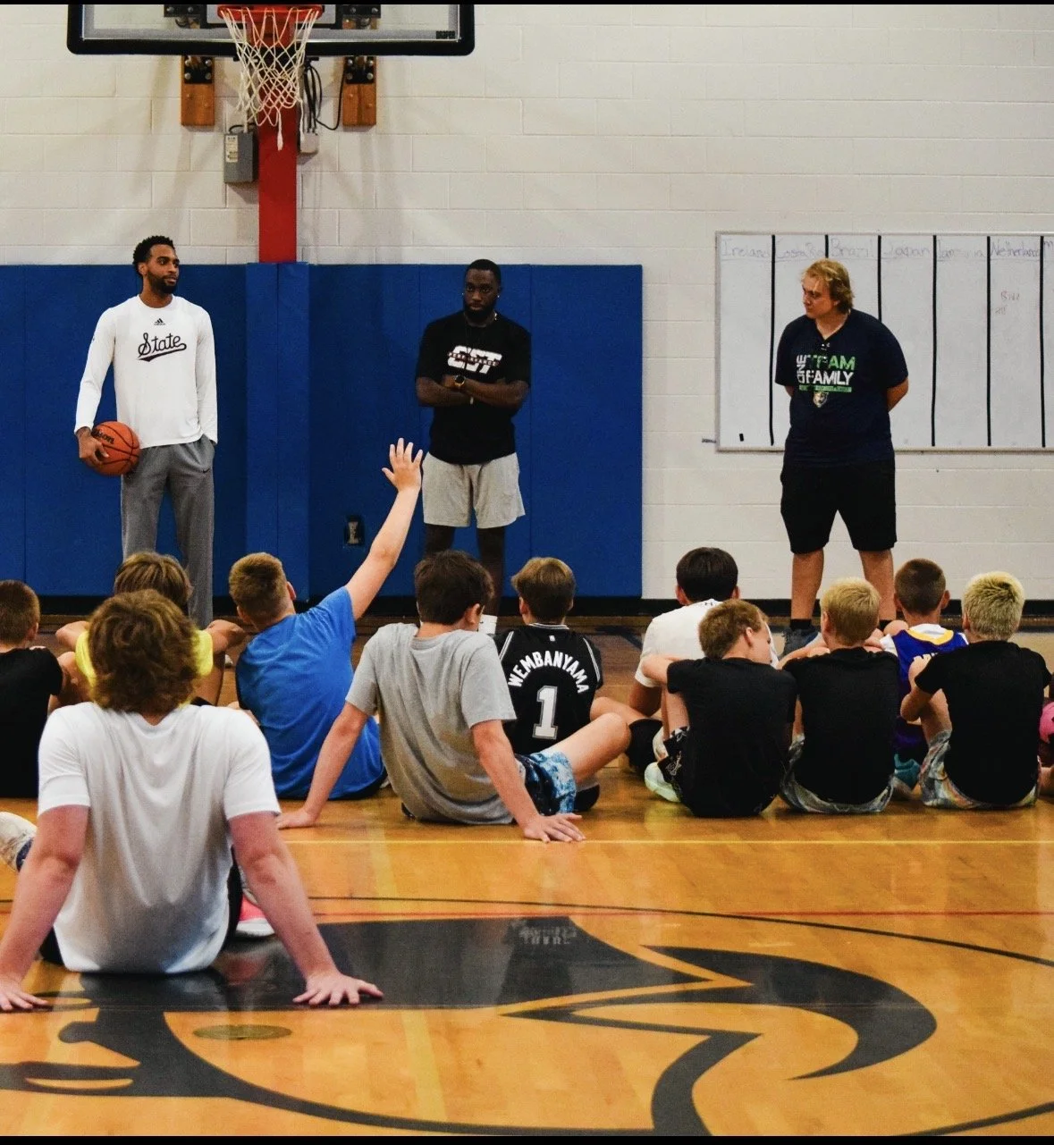Three men standing in front of a group of children sitting on the gym floor during a basketball clinic. One man holds a basketball, another has his arms crossed, and a third stands with hands behind his back. A child sitting on the floor raises their
