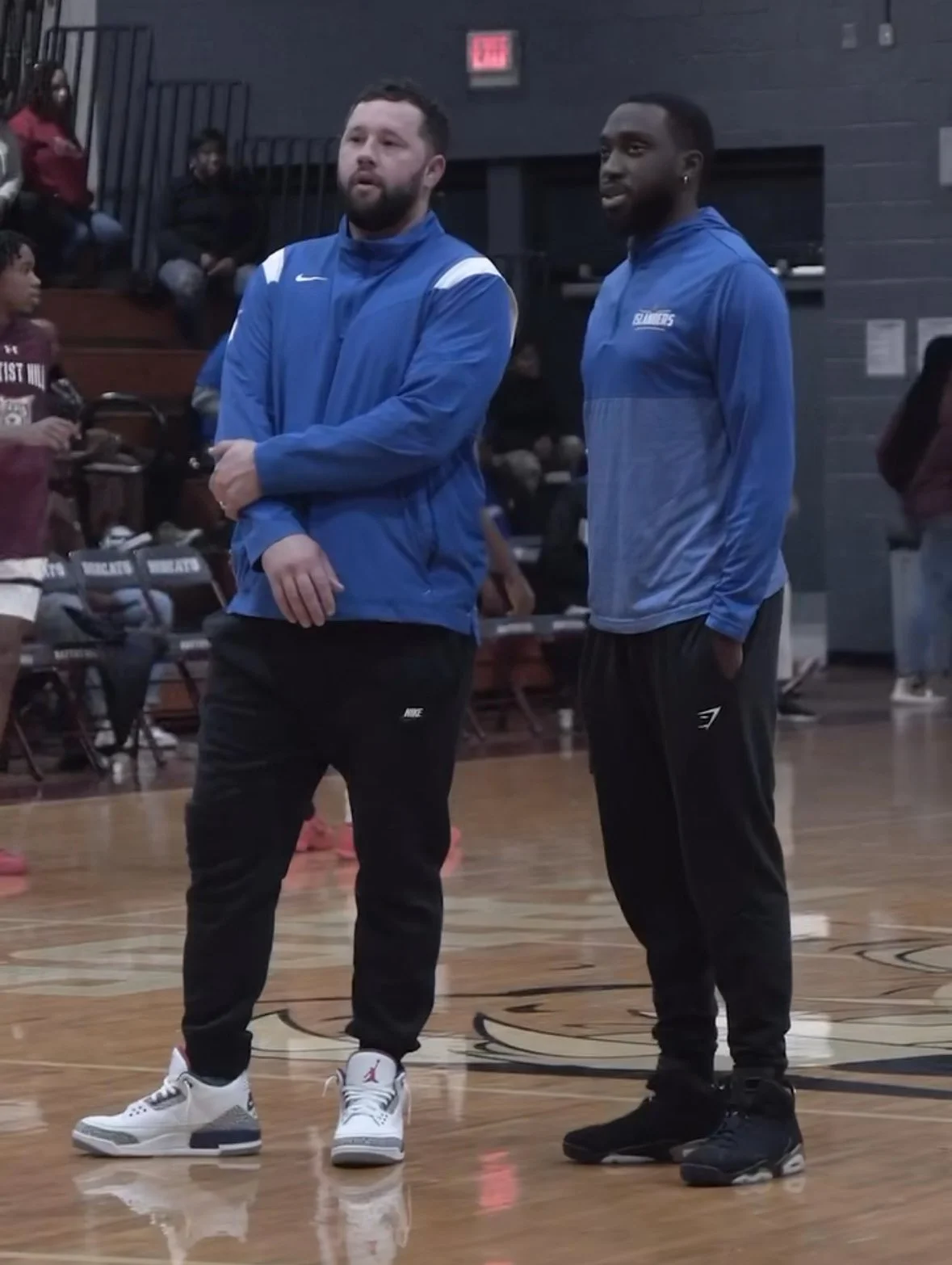 Two men standing on a basketball court in an indoor gym, wearing blue athletic jackets and black pants, engaged in conversation. Several spectators are seated in the background.